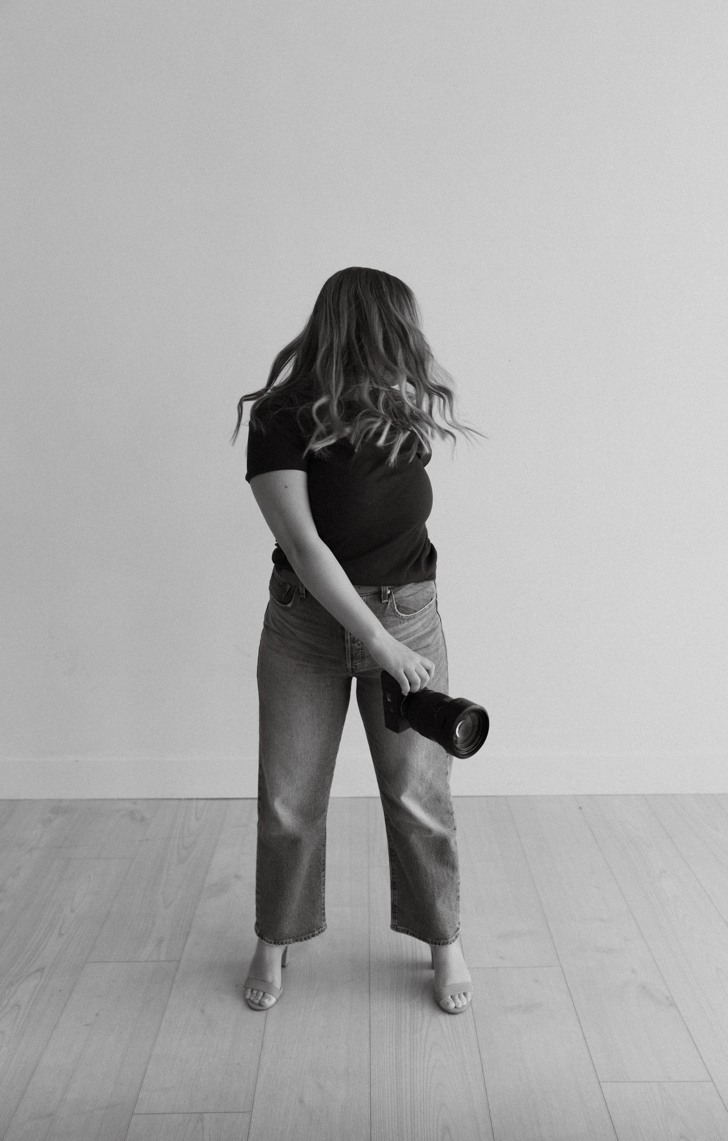 Black and white photo of someone in casual wear holding camera against white wall.