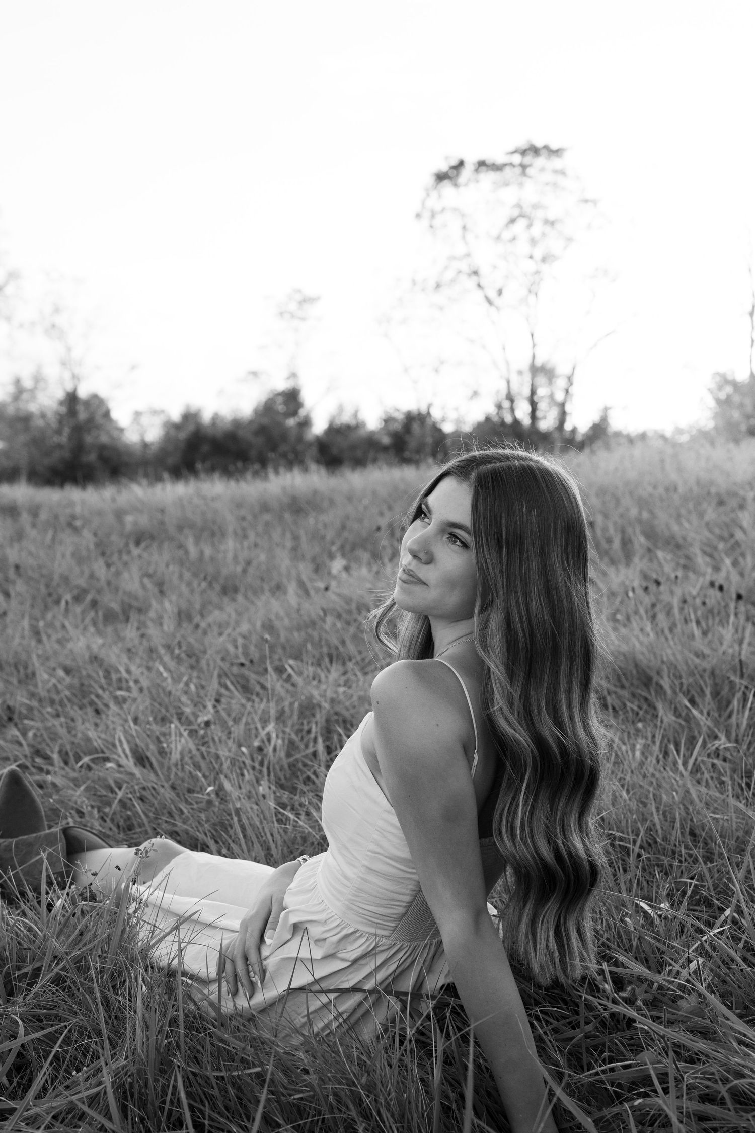 Black and white photo of a young woman with long wavy hair sitting in a grassy field, wearing a white dress.