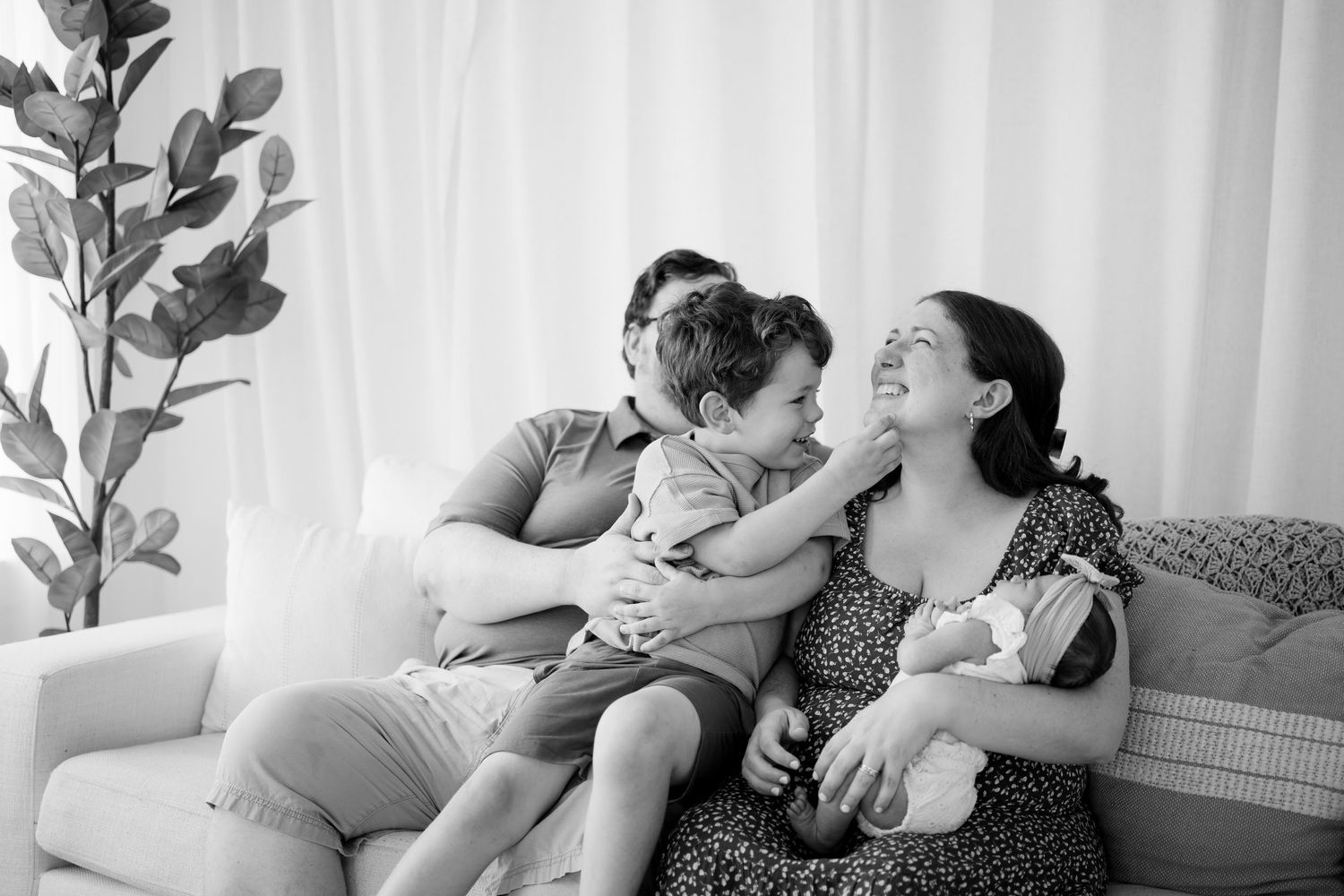 Black and white photo of a happy family with two parents, a newborn, and a toddler laughing together on a cozy sofa in Novi, Michigan.