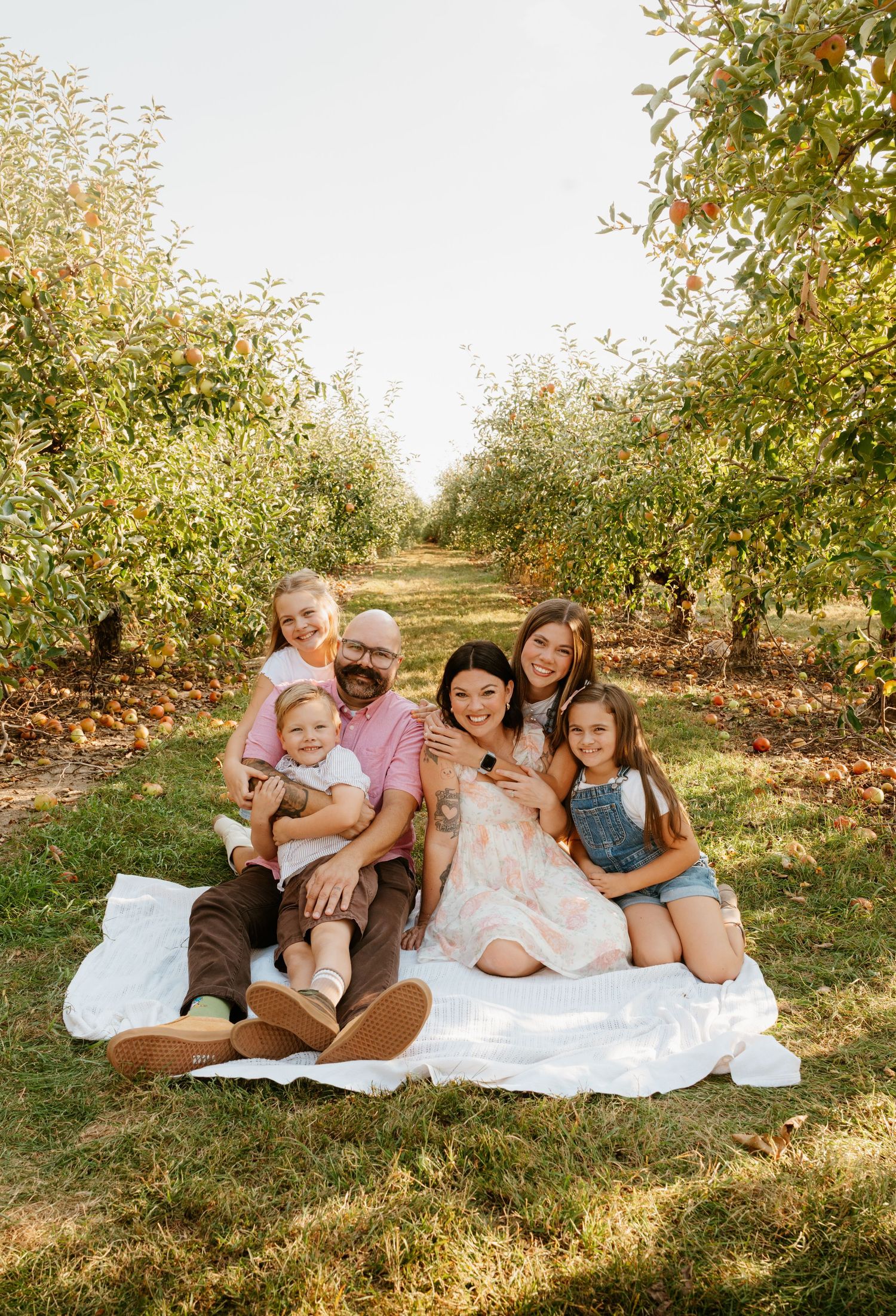 Family group relaxing on a white blanket under apple trees in an orchard.