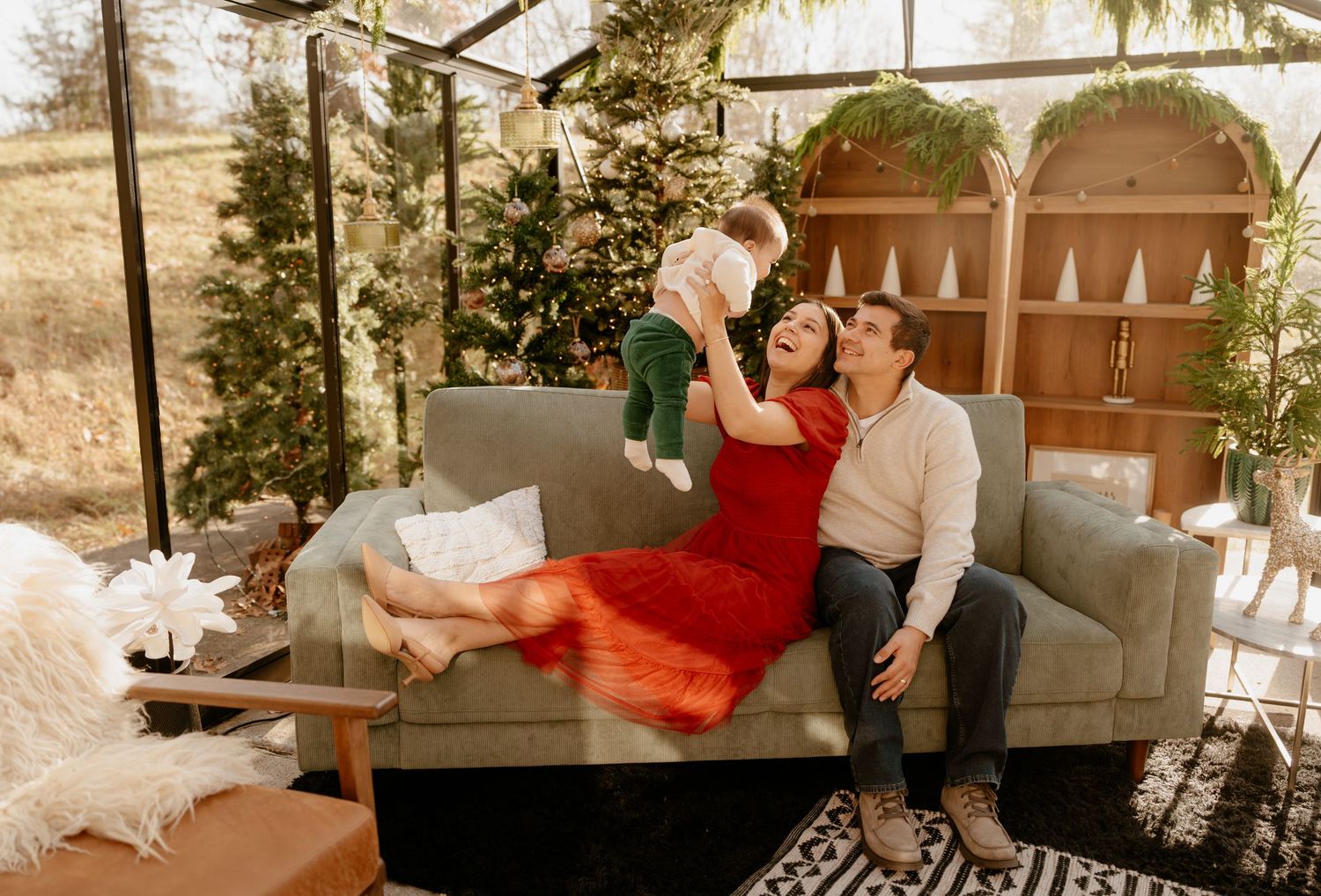 Family holiday photo with mother in red dress lifting baby, father seated on gray sofa, Christmas trees in background.