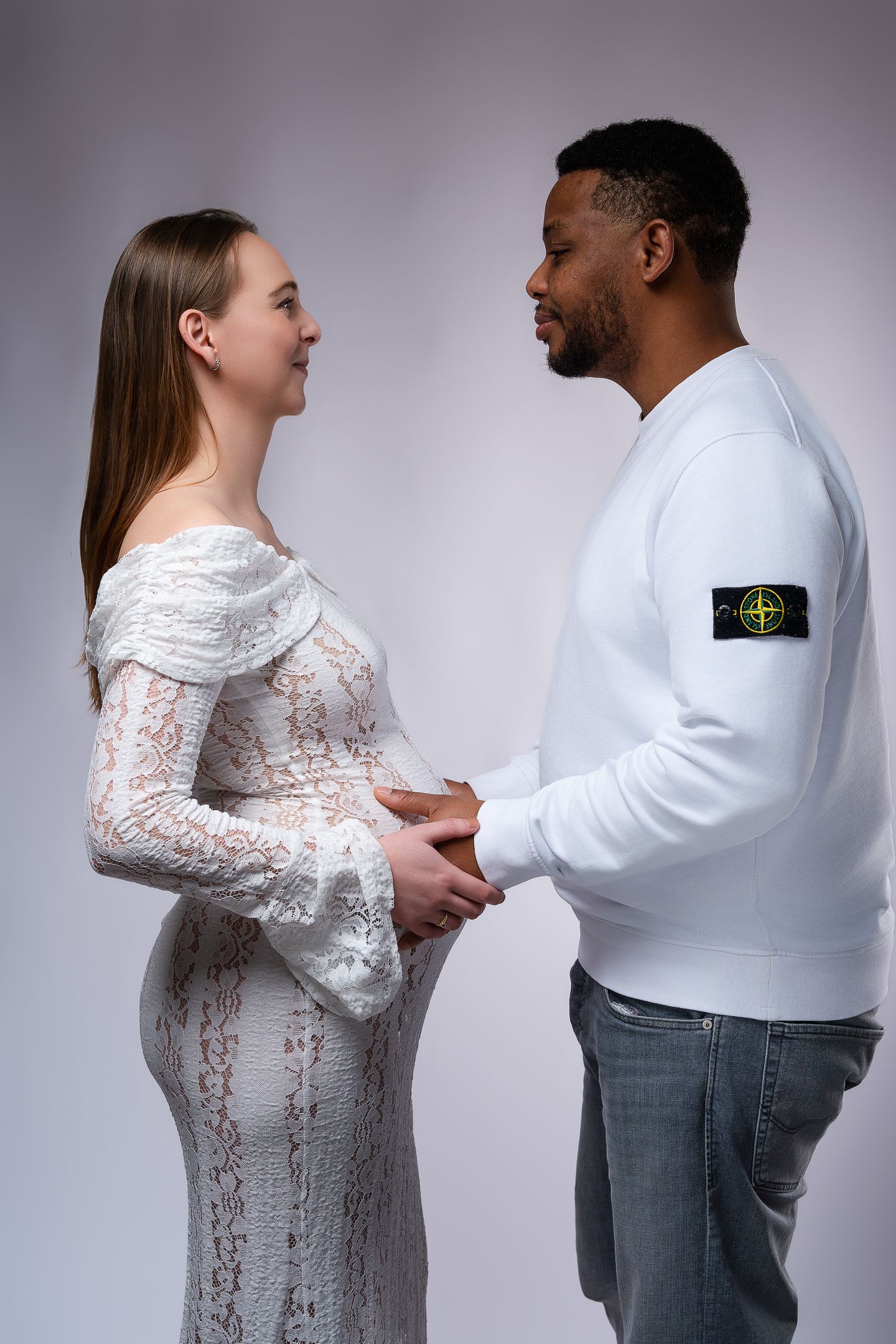 A pregnant woman in a white lace dress holds hands with a man in a white Stone Island sweatshirt and jeans.