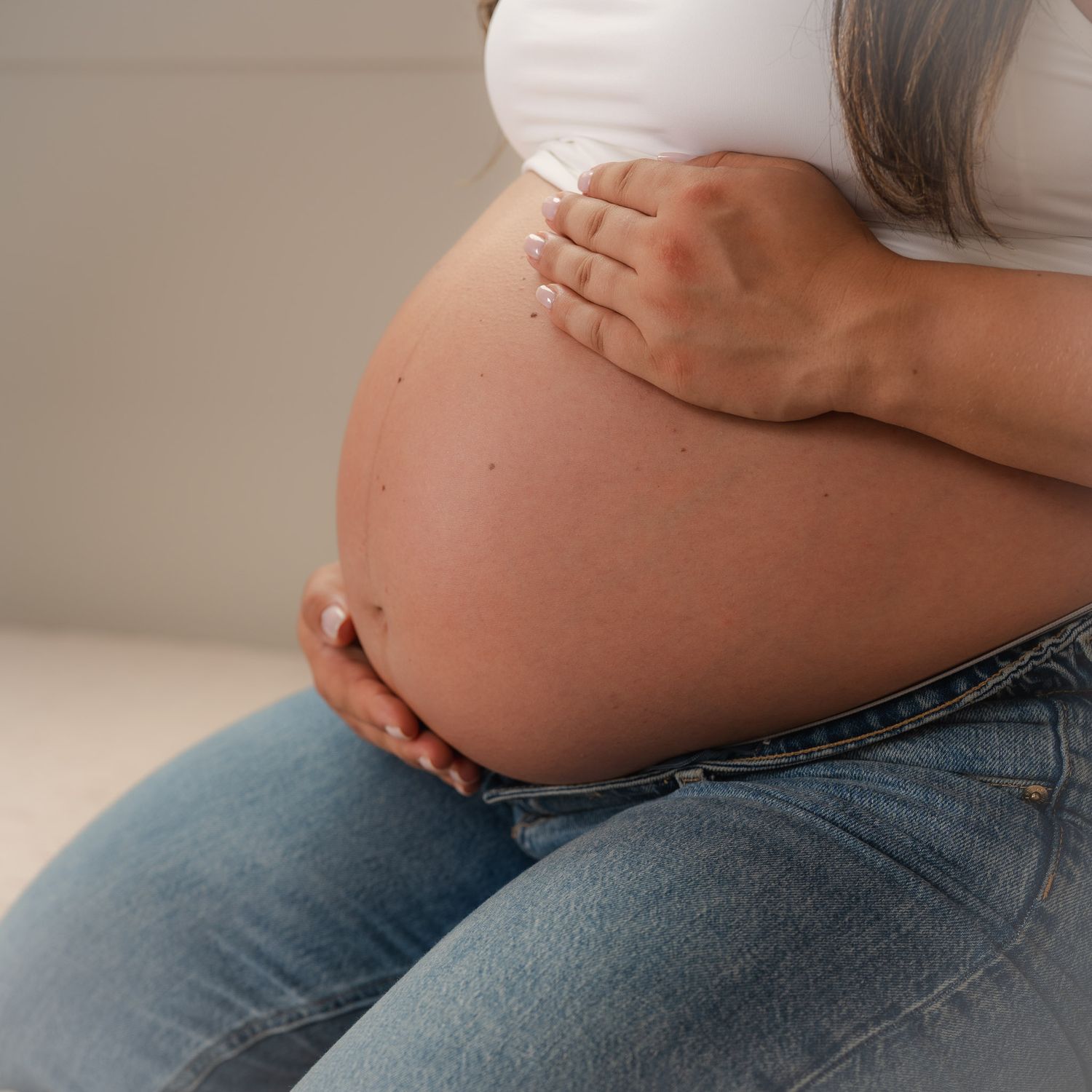 Pregnant woman in jeans cradling her bare baby bump with both hands, wearing a white top.