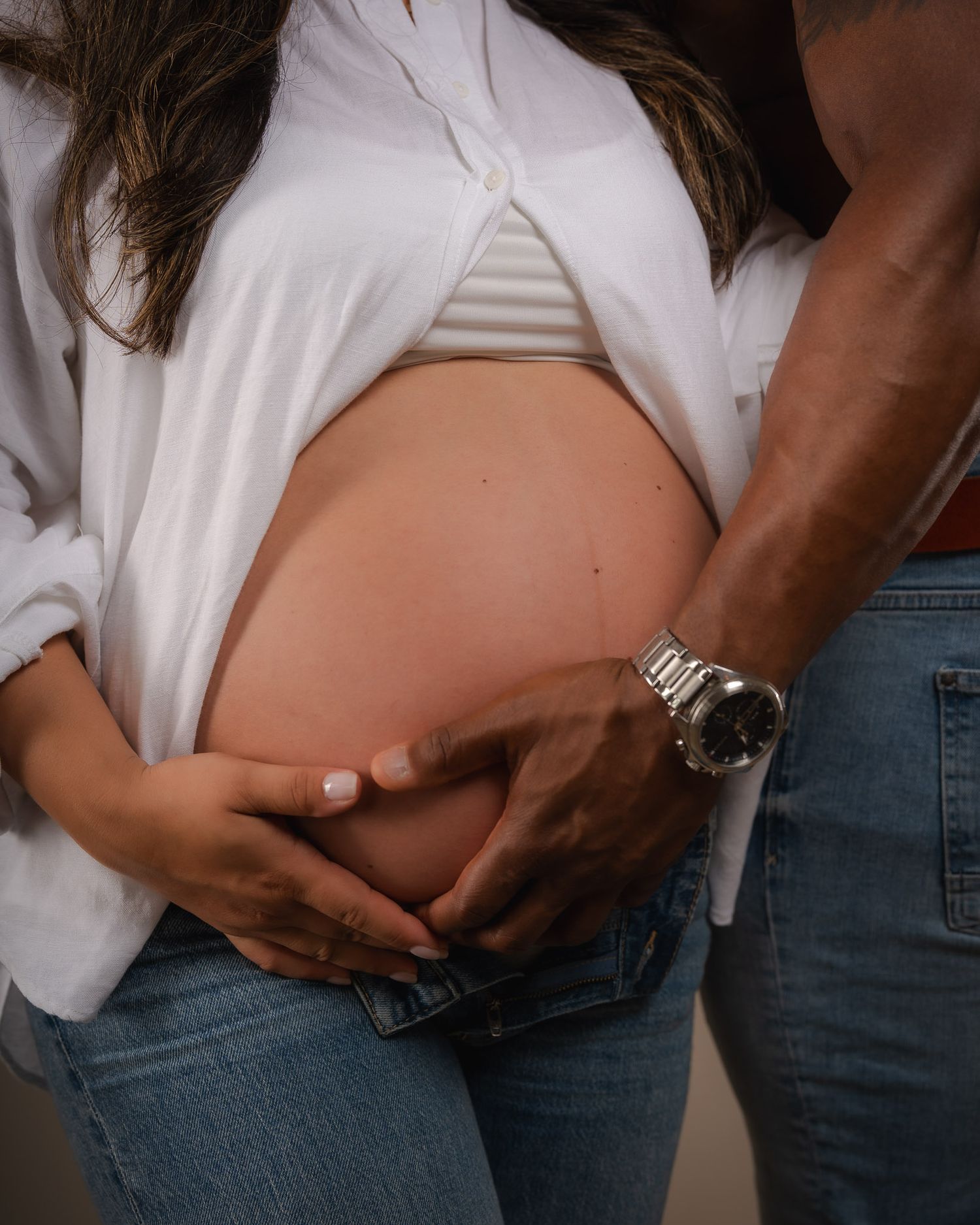 Couple tenderly cradling pregnant belly, woman in white shirt and jeans, partner wearing silver watch.