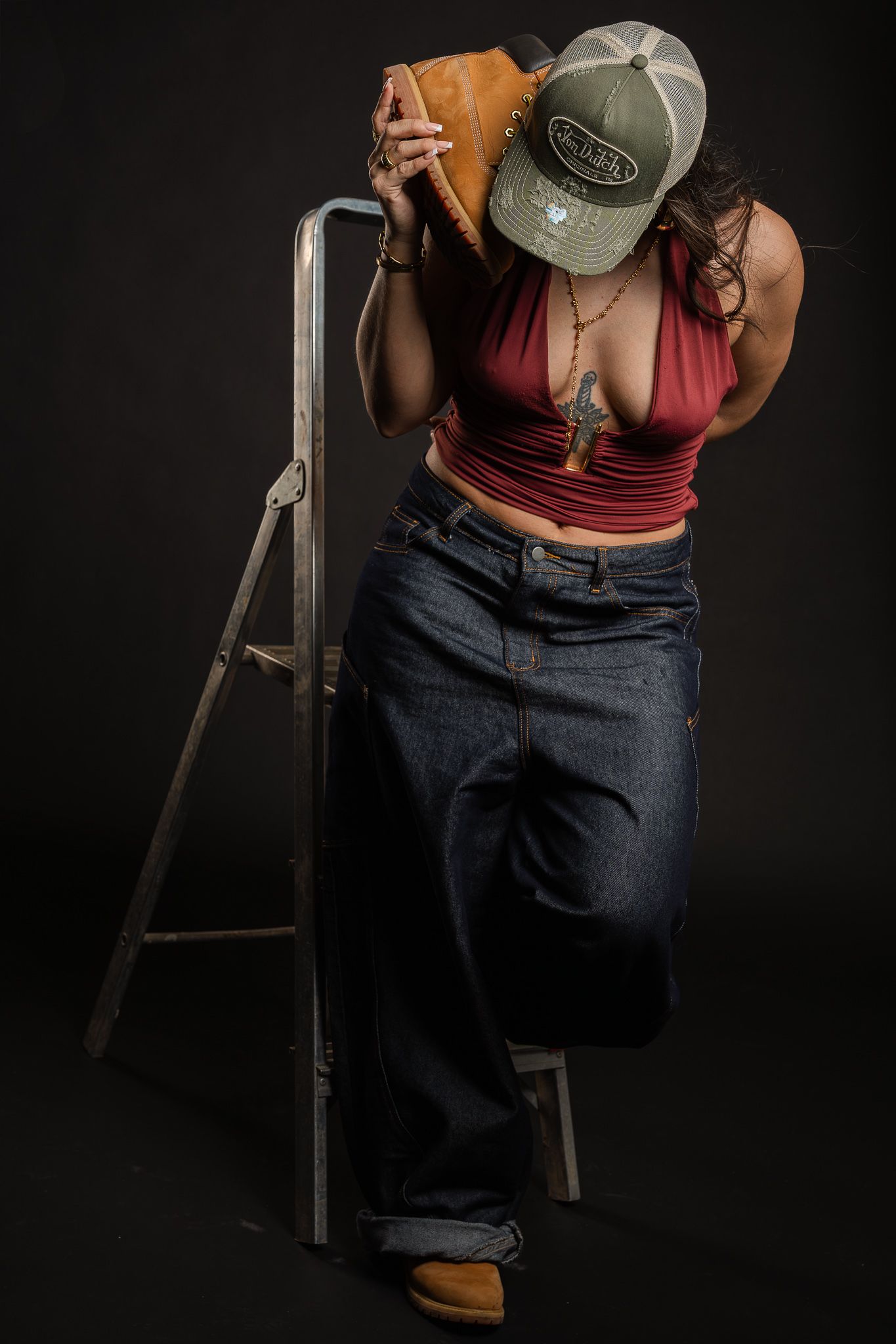 Person wearing a gray baseball cap, burgundy tank top and dark jeans sits on a wooden ladder against a black background.