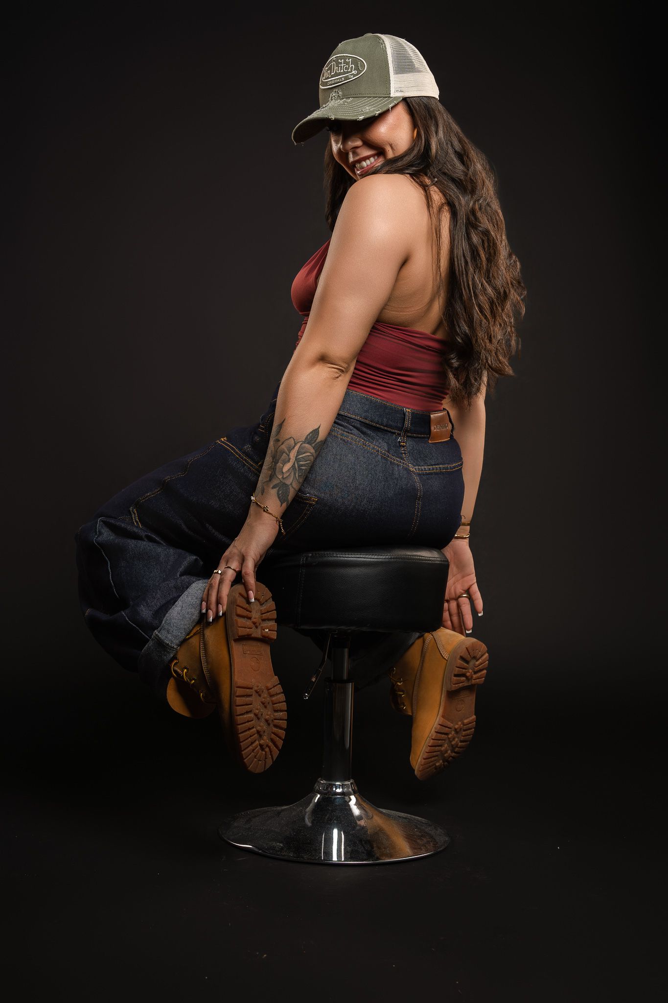 Woman in burgundy top and jeans poses on reflective stool against dark background wearing brown boots.