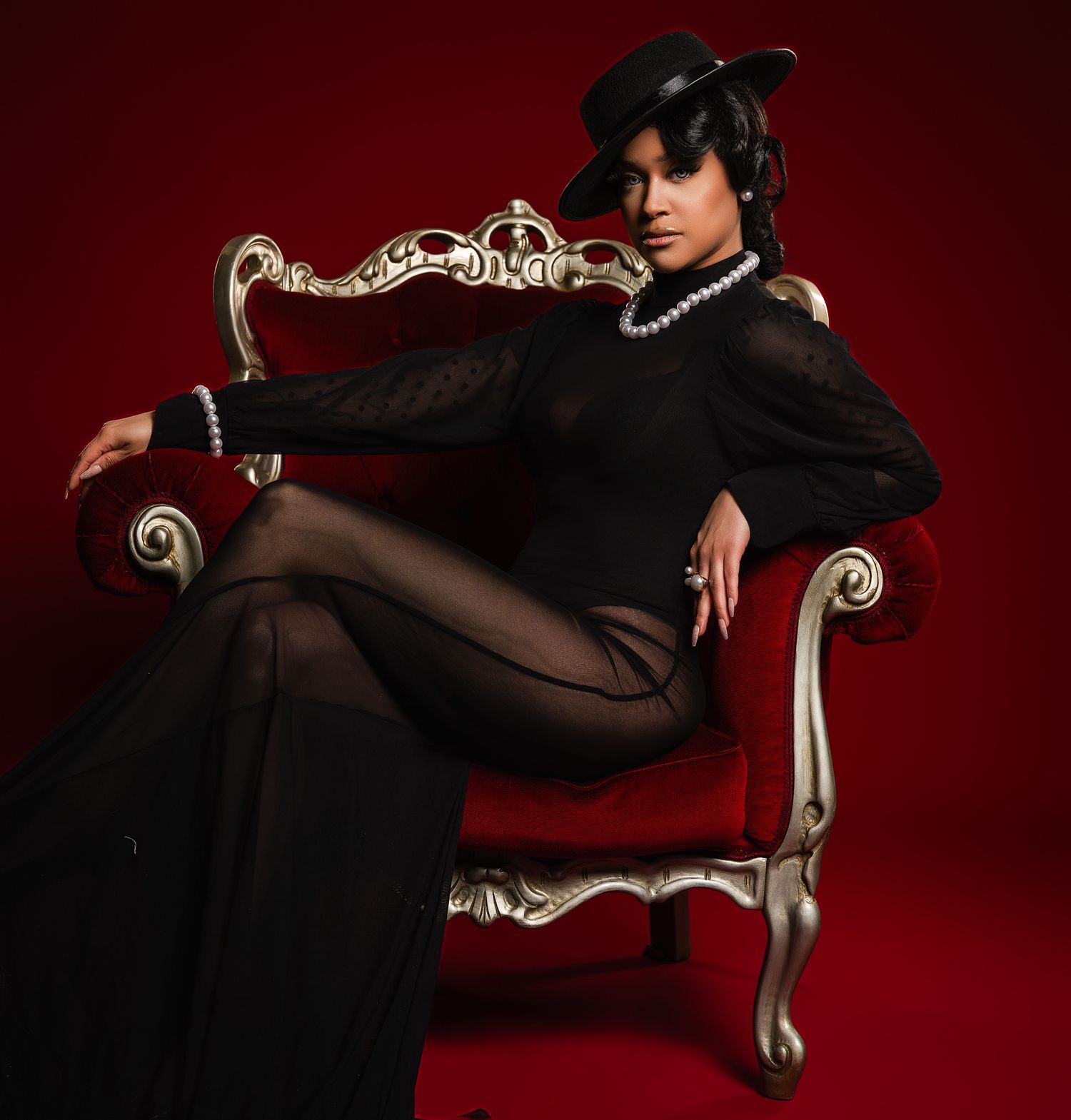 A dramatic portrait on an ornate silver chair against a deep red background features black clothing and a stylish hat.