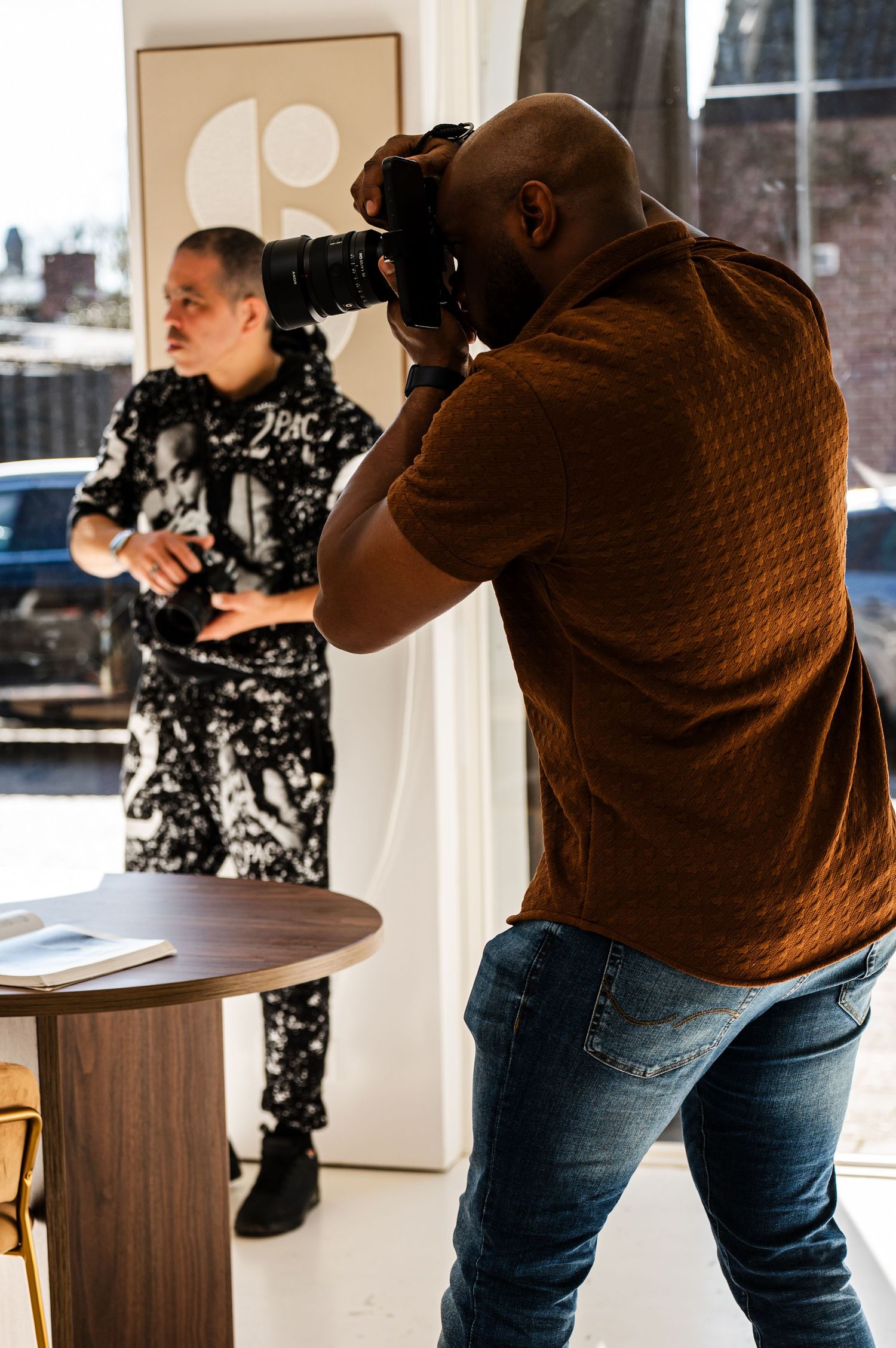 A photographer in a brown shirt takes pictures while another person in patterned clothing stands nearby in an indoor setting.