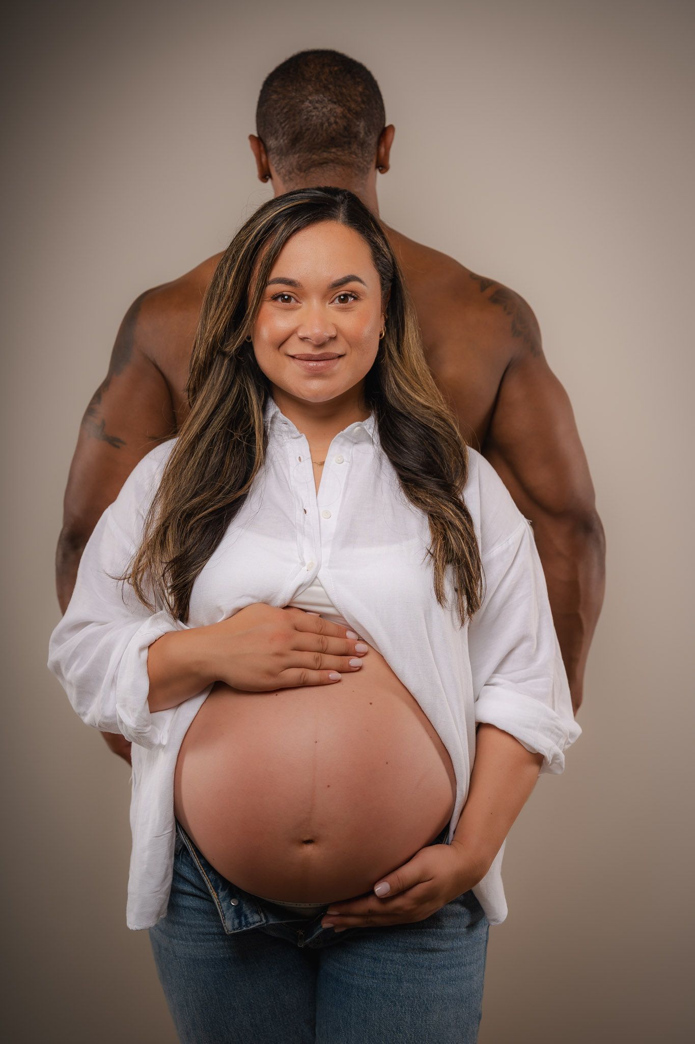 A pregnant woman in a white shirt holds her bare belly while a shirtless man stands behind her in a studio.