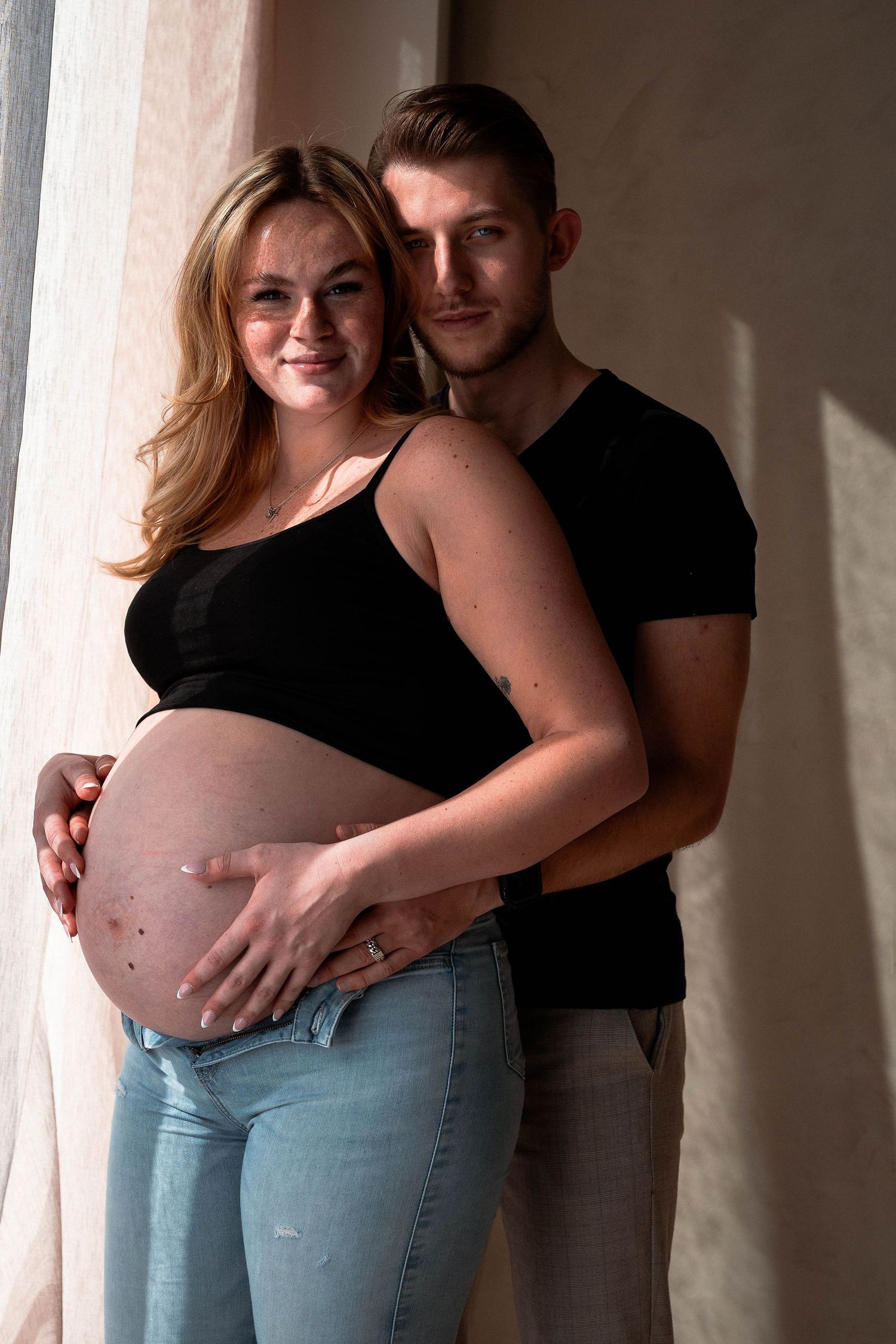 A series of intimate maternity photos showing a couple embracing in casual attire against a neutral wall.