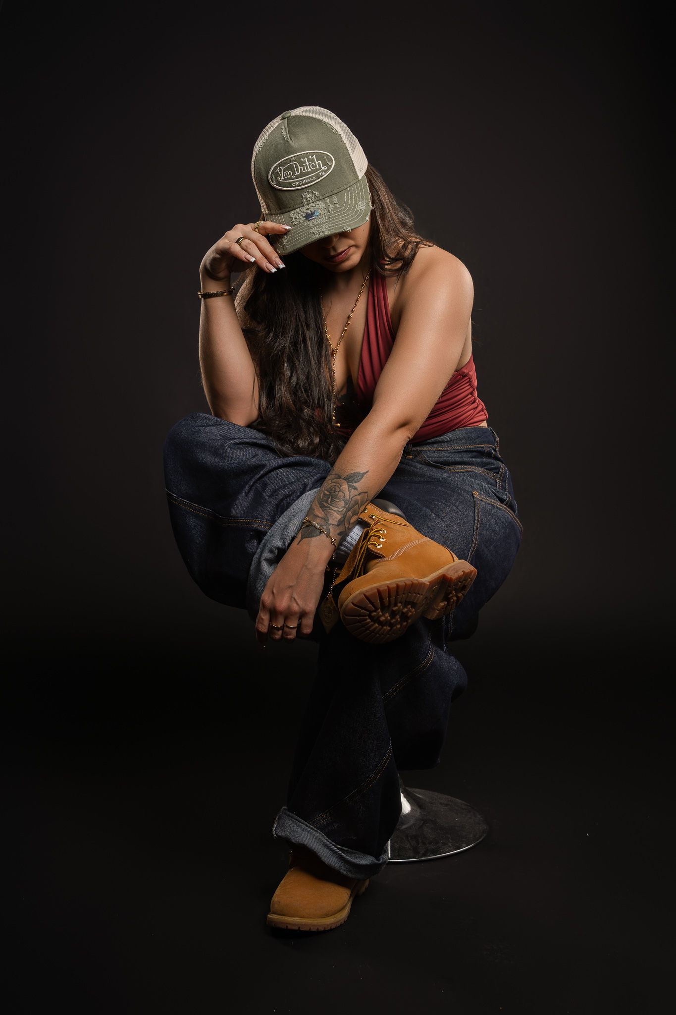 Person wearing a baseball cap and burgundy tank top crouches in a contemplative pose against a dark background.