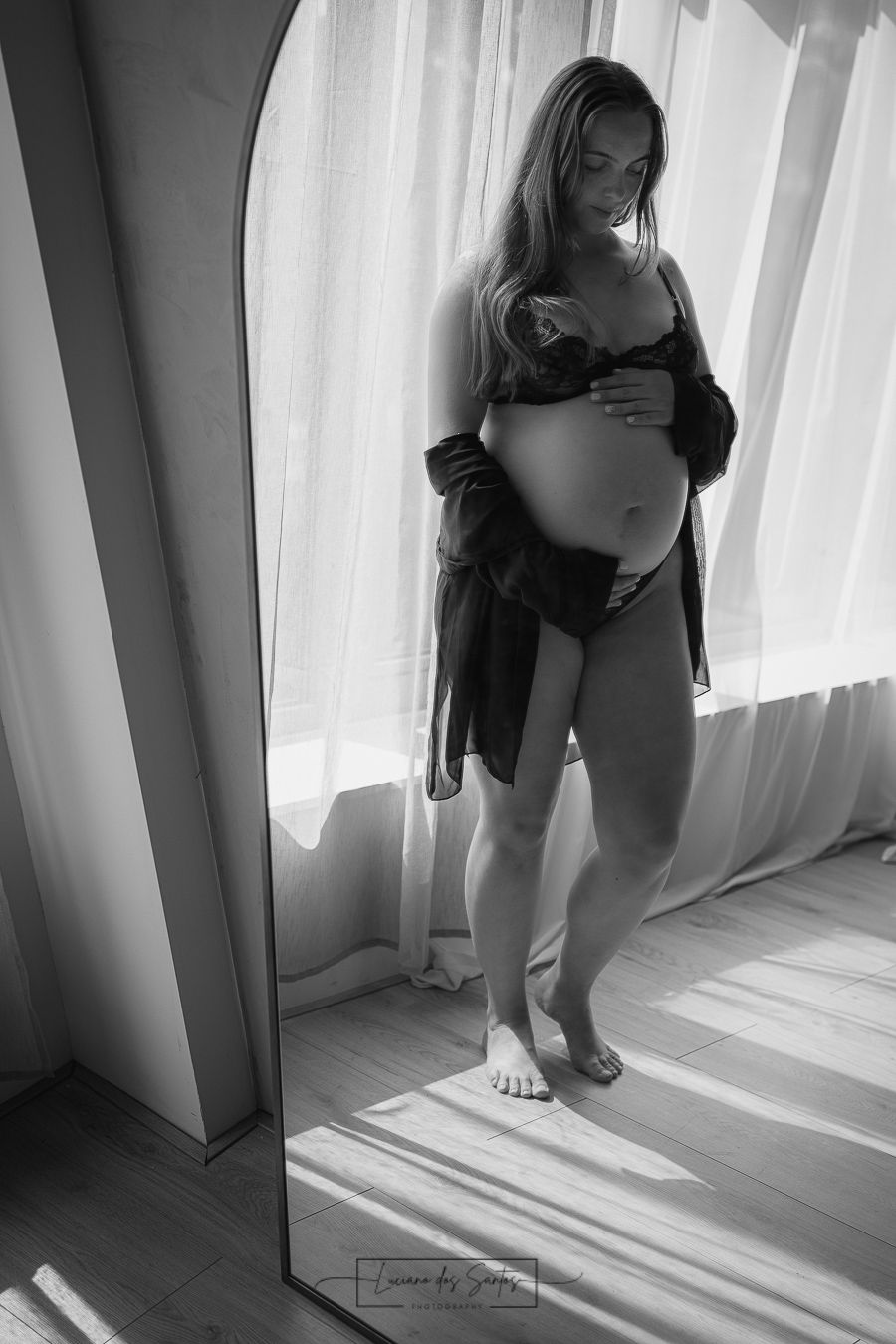 Black and white artistic maternity photo with natural window light casting shadows on wooden floor.