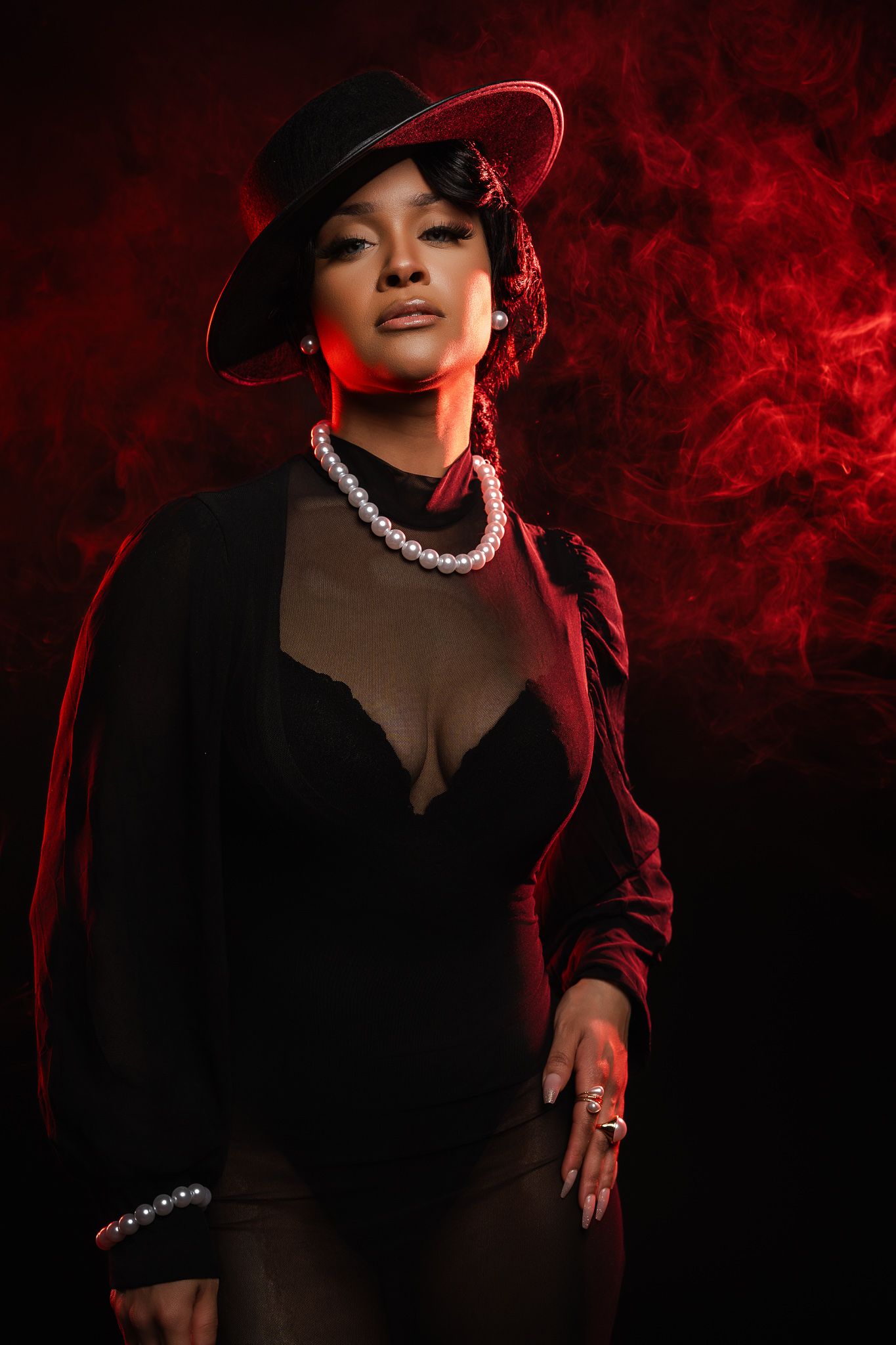 Dramatic portrait of figure in black dress and hat with pearl necklace against red smoky background.