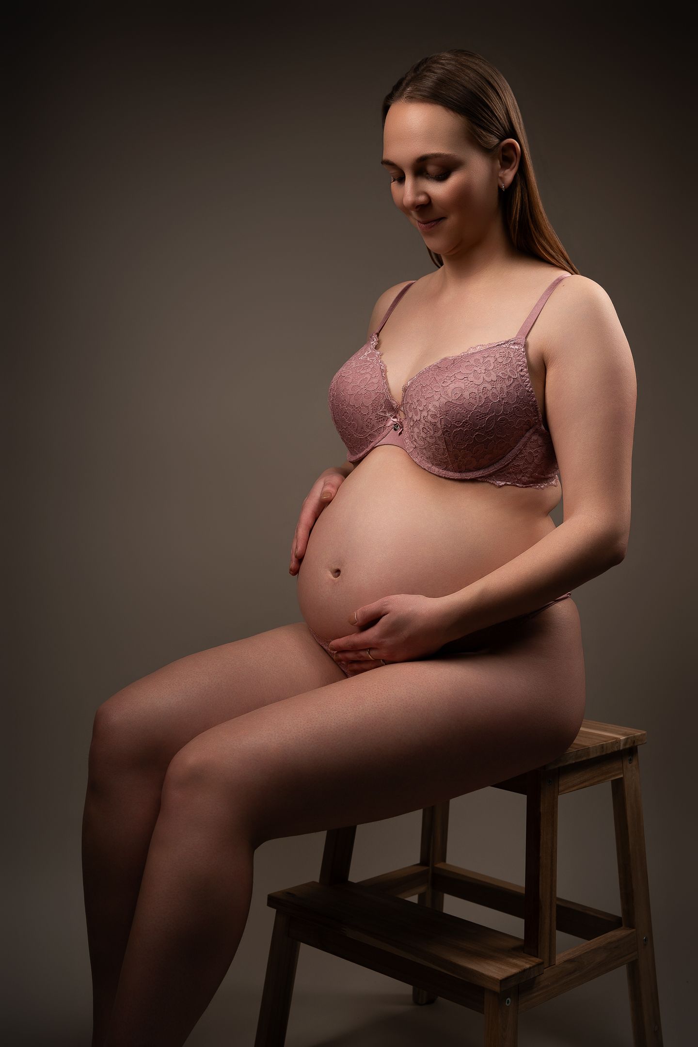 Pregnant woman in pink lace bra sitting on wooden stool, cradling baby bump, studio portrait on gray background.