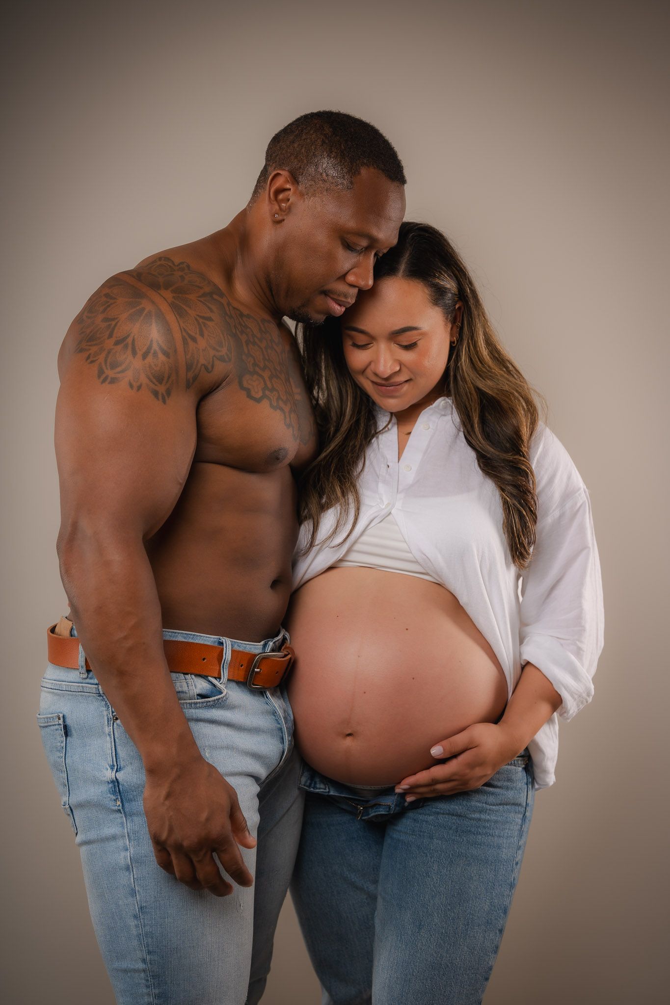 A shirtless tattooed man and a smiling pregnant woman in jeans pose together, cradling her baby bump in a studio.