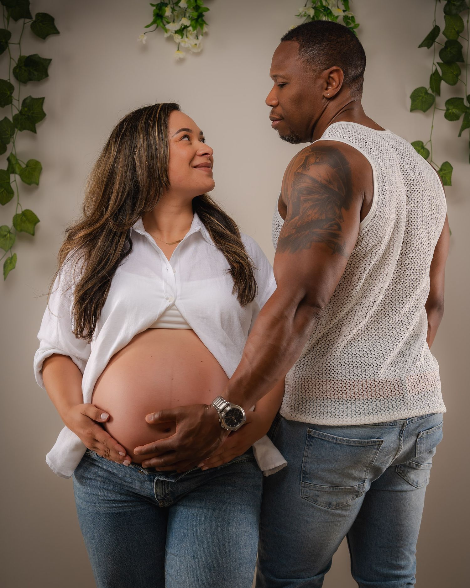 Couple sharing a tender maternity photo shoot moment, pregnant woman in white shirt and jeans with partner holding her baby bump.