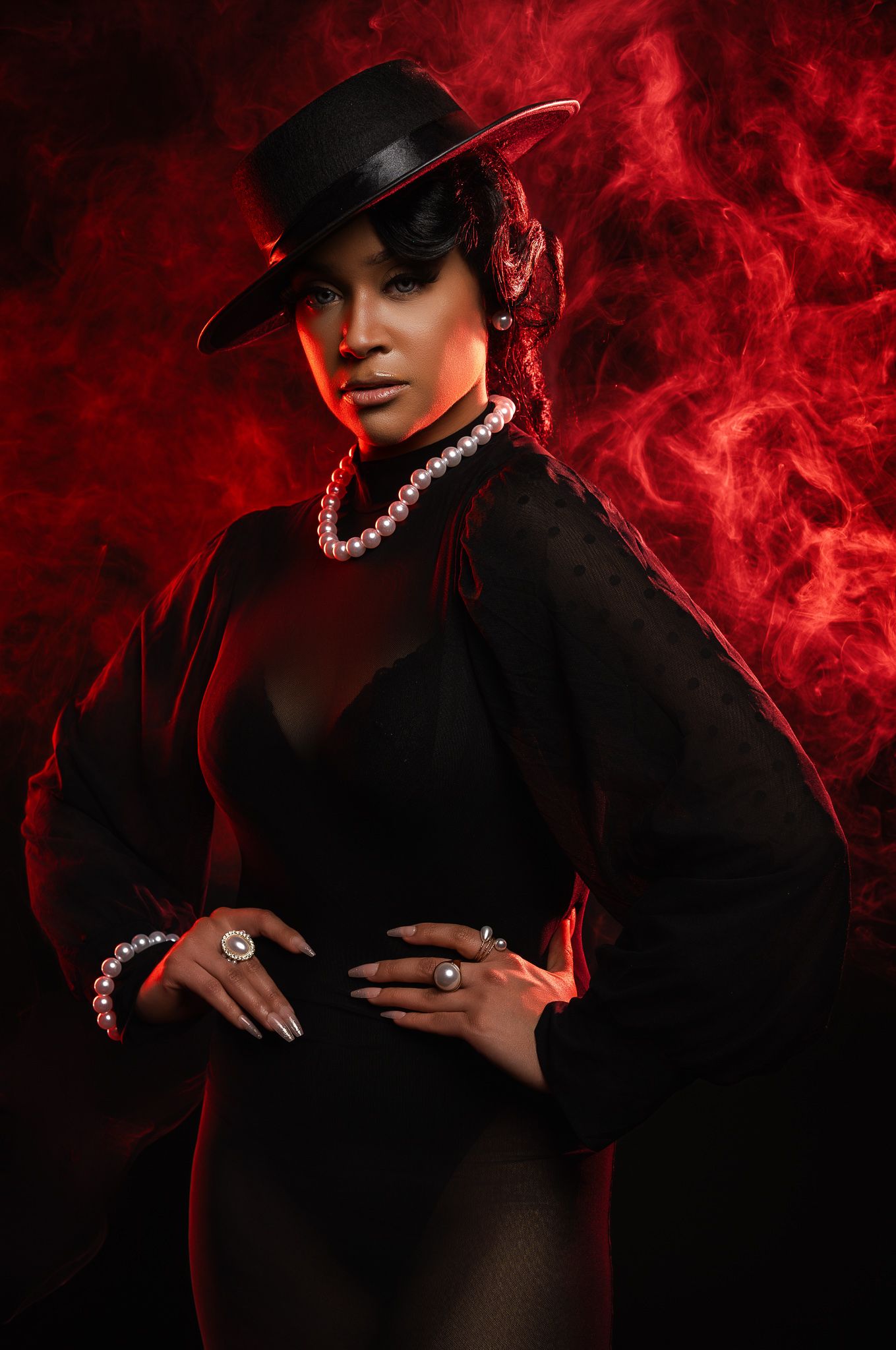 Dramatic fashion portrait with black dress, hat and pearl necklace against swirling red smoke background.