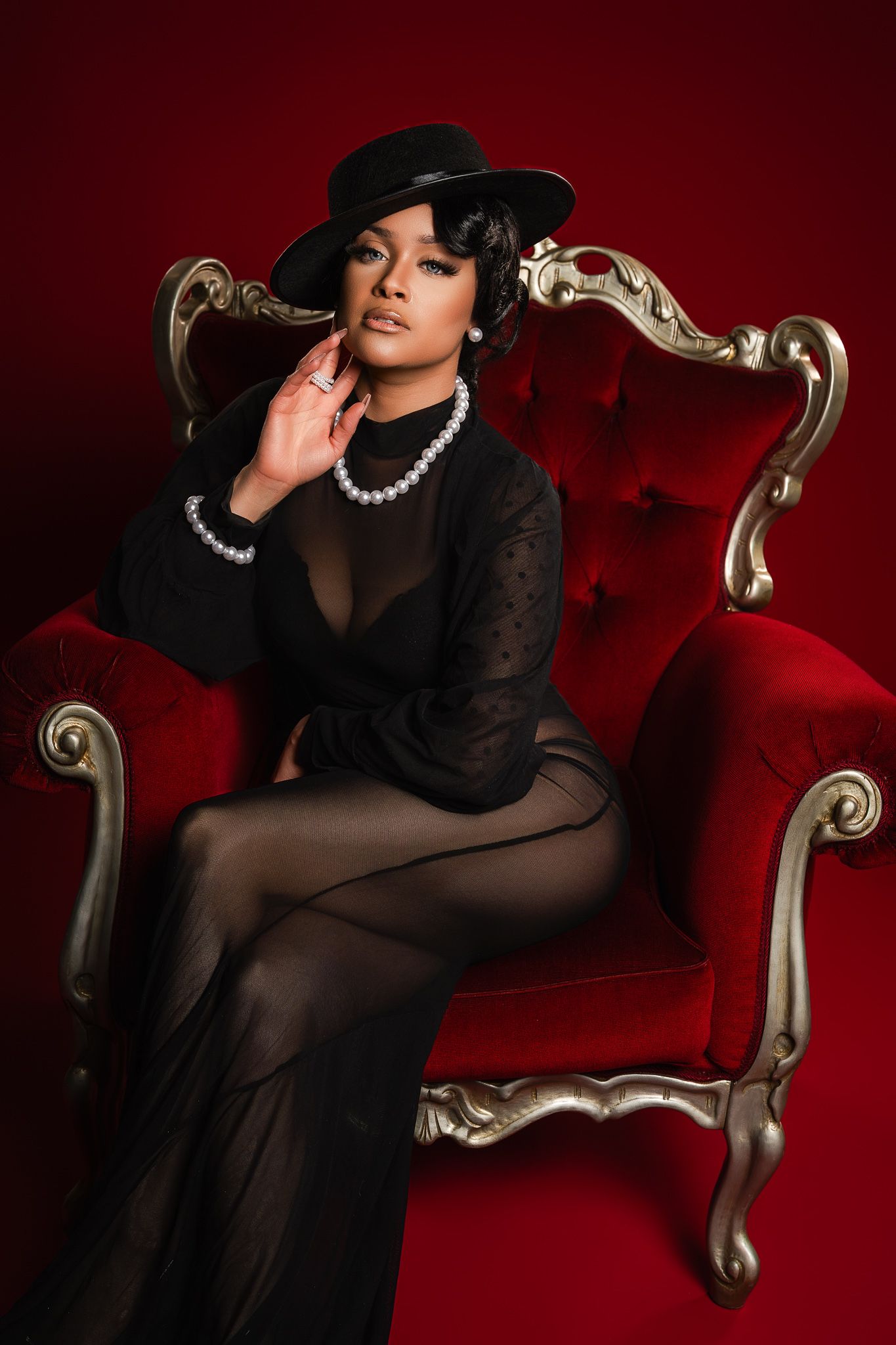 Dramatic fashion portraits against red backdrop show figure in black sheer dress and hat posing on ornate vintage chair.