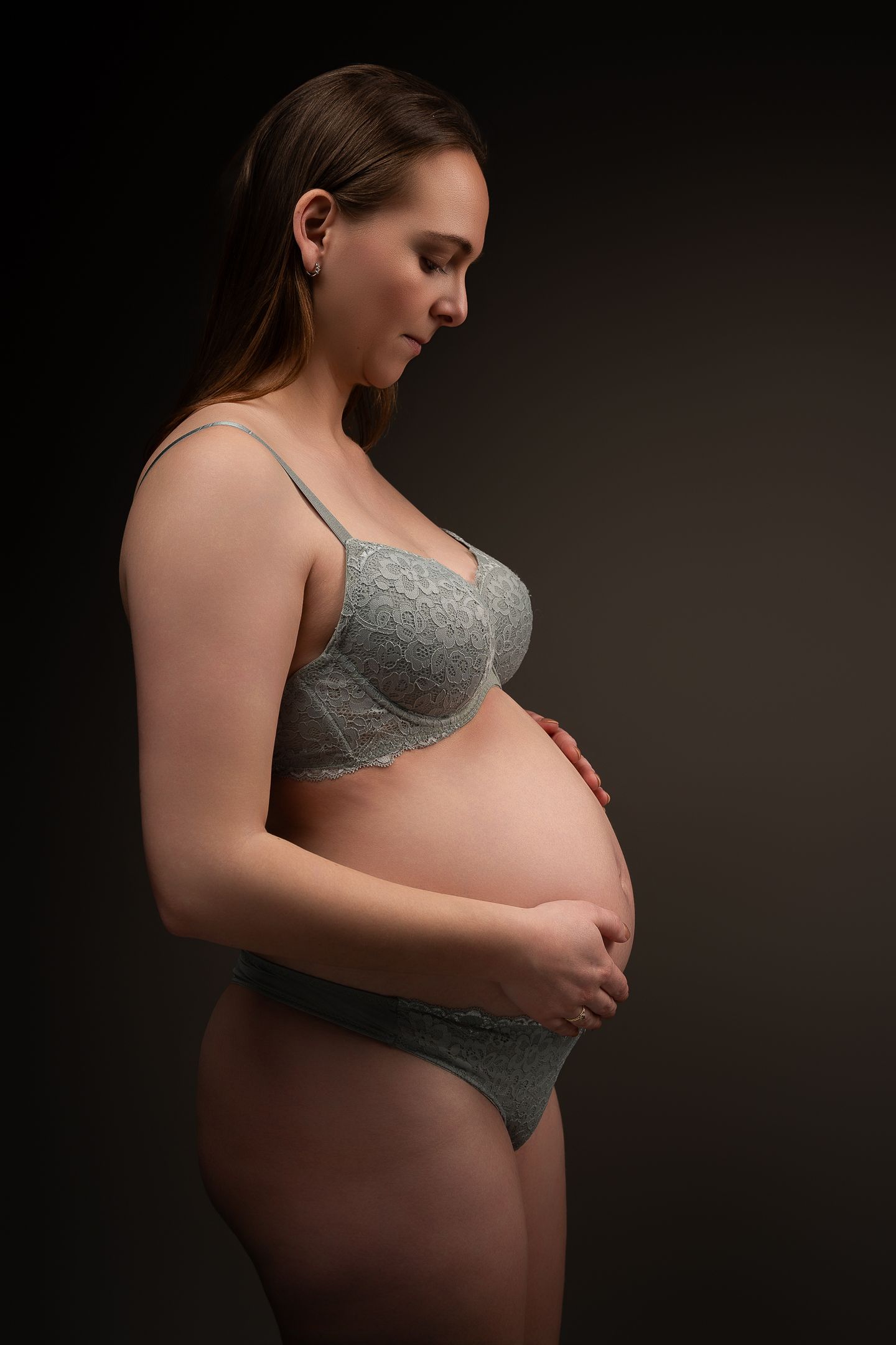 Pregnant woman in gray lace lingerie cradling her baby bump against a dark background.