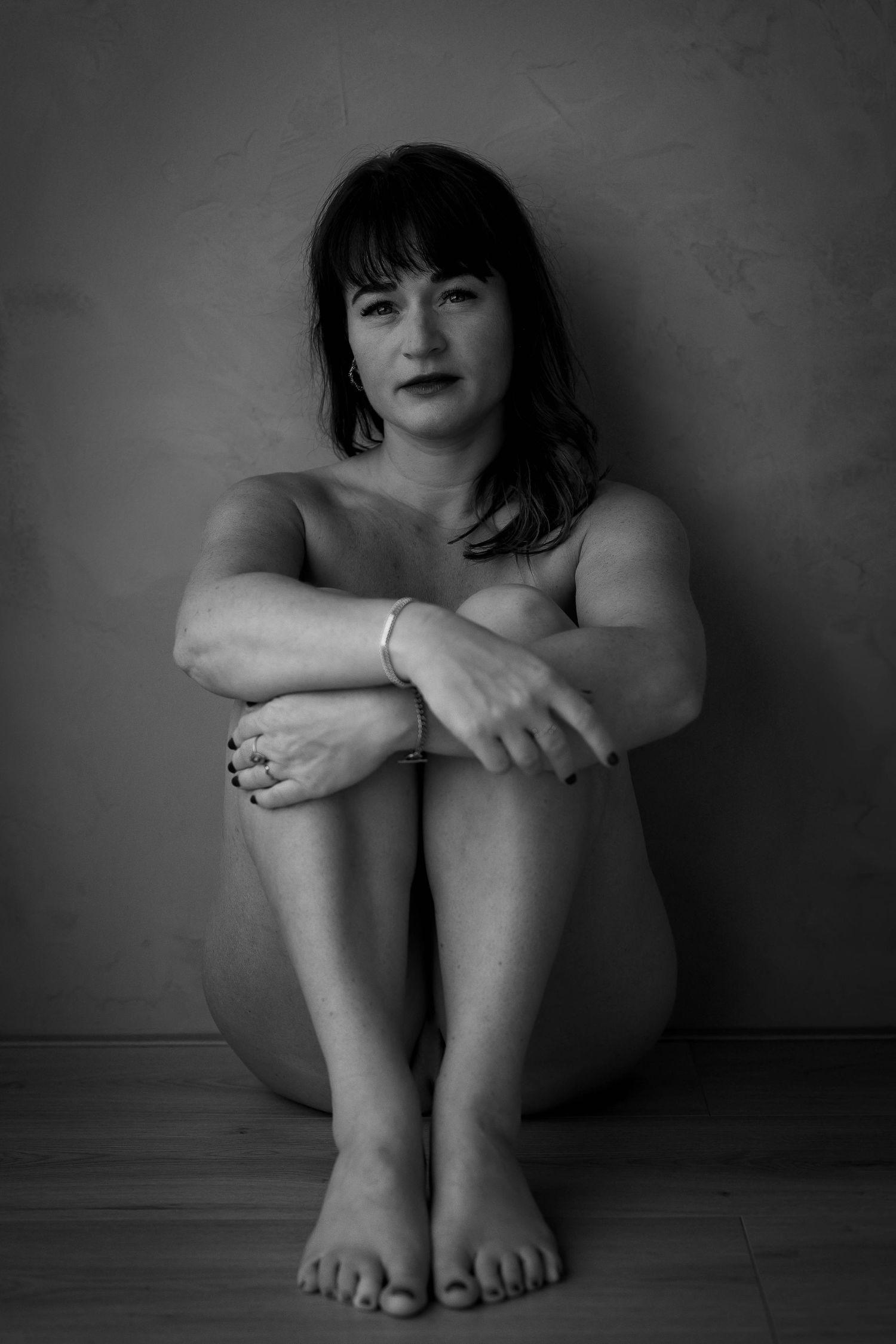 Black and white artistic portrait of someone sitting with knees drawn up, wearing dark lipstick and bangs.