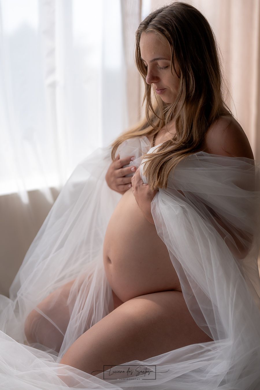 Ethereal maternity photo with flowing tulle fabric in window light.