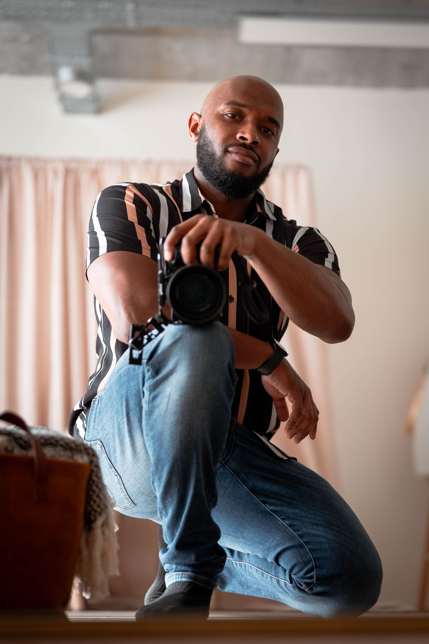 Photographer in striped shirt and jeans crouches while holding camera in indoor setting with soft lighting.