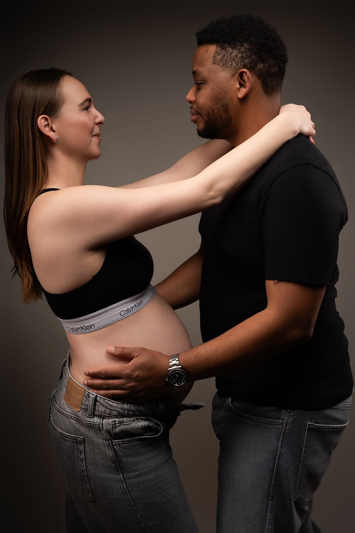 A pregnant woman in a Calvin Klein crop top embraces her partner, both smiling warmly in a studio setting.