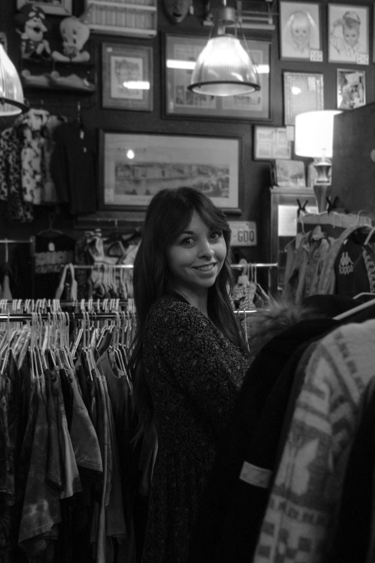 A smiling woman standing among clothing racks in a cozy vintage boutique, black and white.