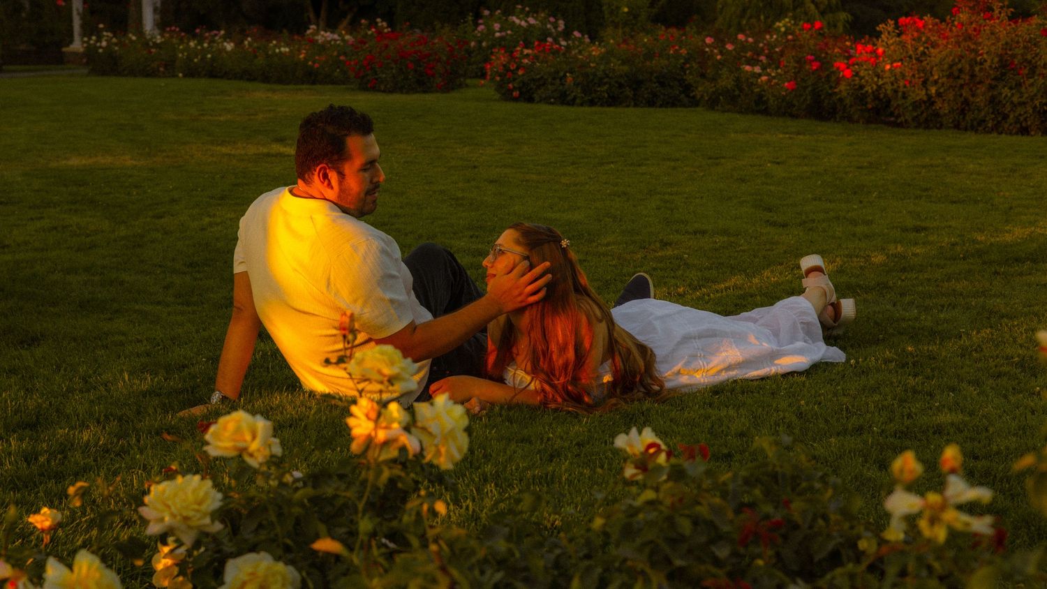 Romantic couple relaxing on lush green grass surrounded by blooming flowers in a garden at golden hour.