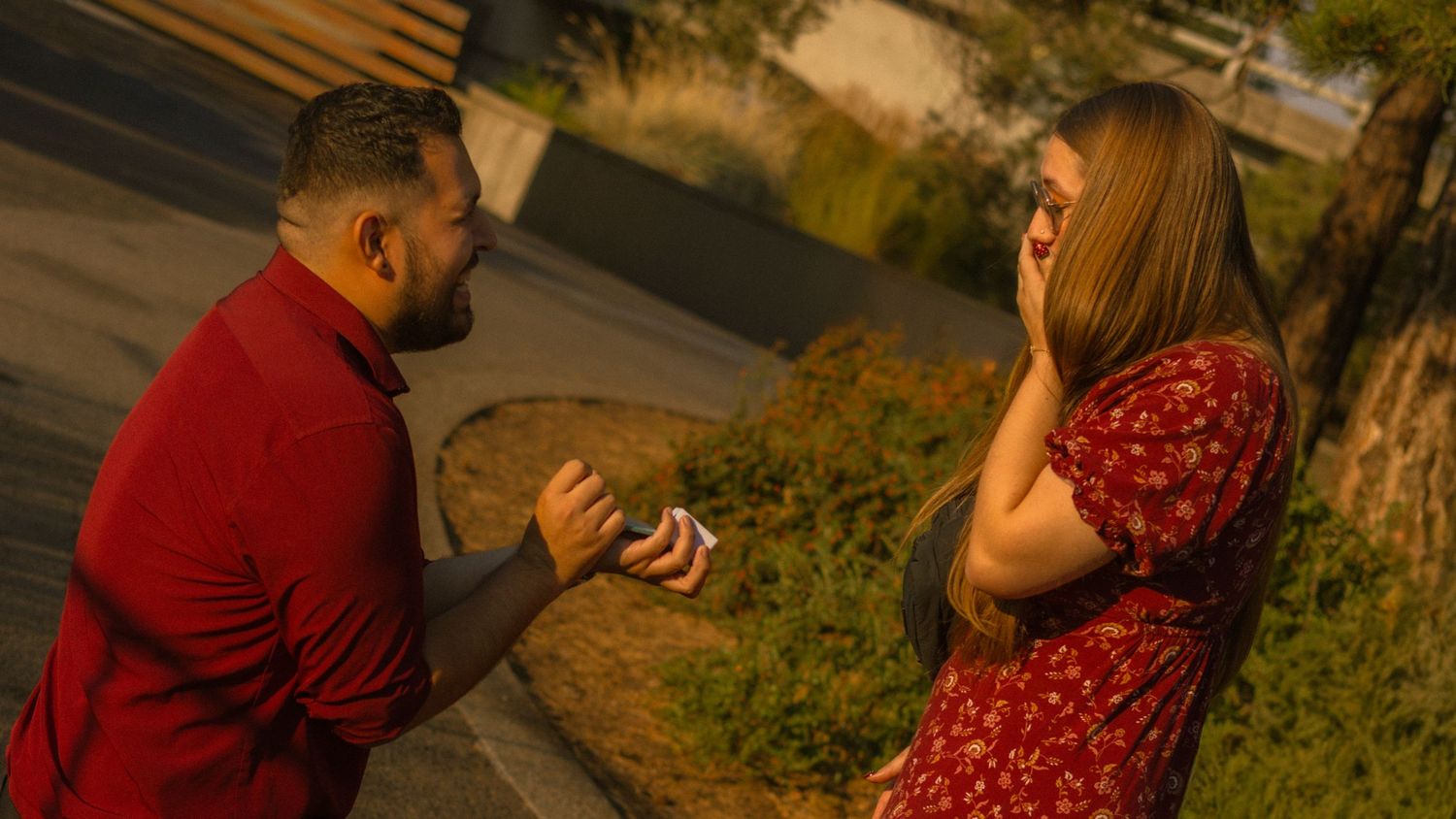 Man in red shirt proposes to surprised woman in floral dress outdoors during golden hour.
