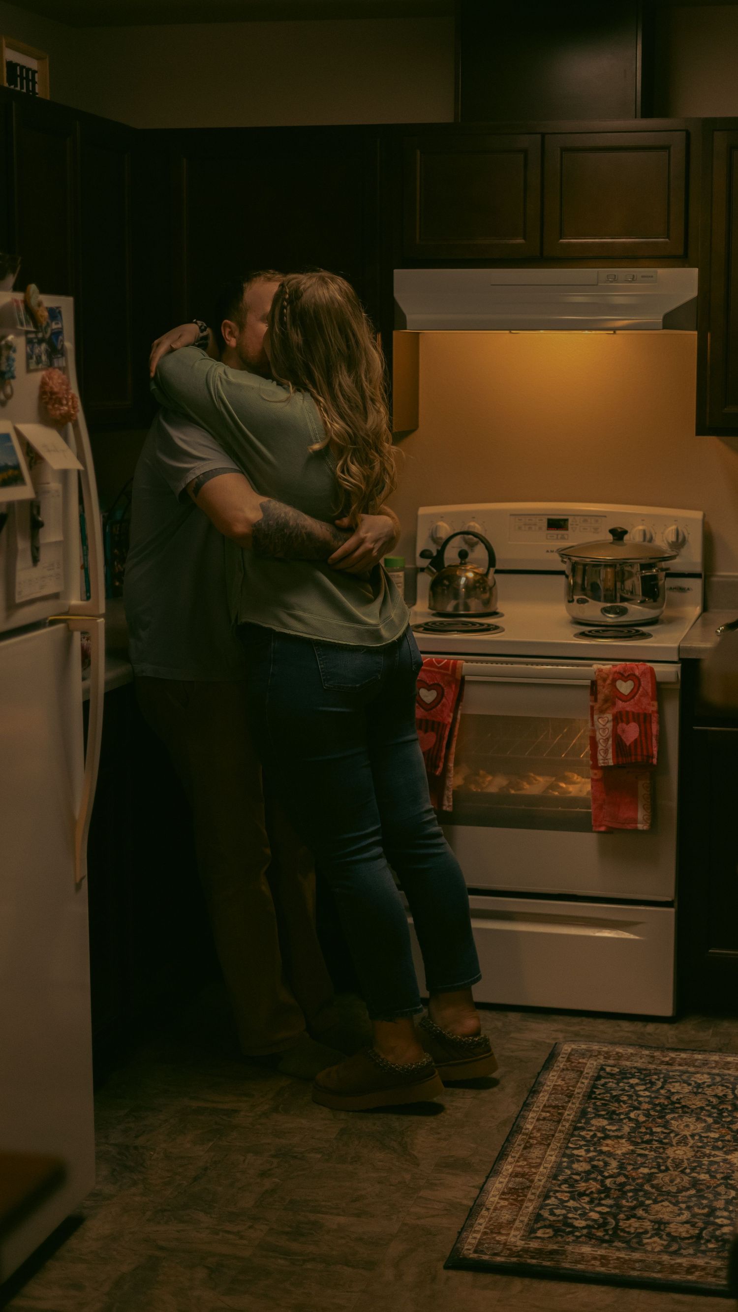Couple sharing a tender embrace and kiss in a warmly lit kitchen near the stove.