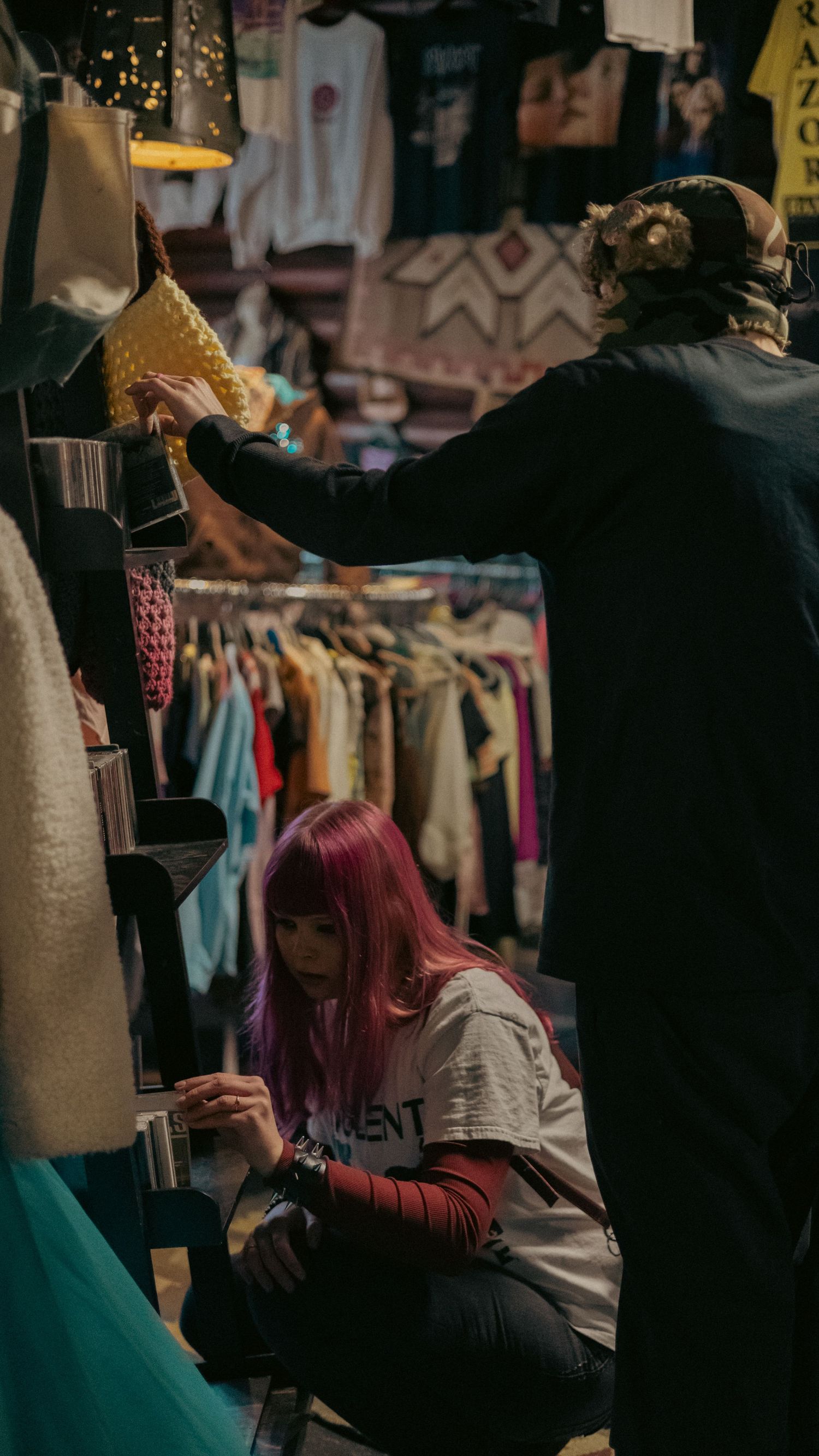 A woman with pink hair crouching while shopping in a colorful vintage clothing store.
