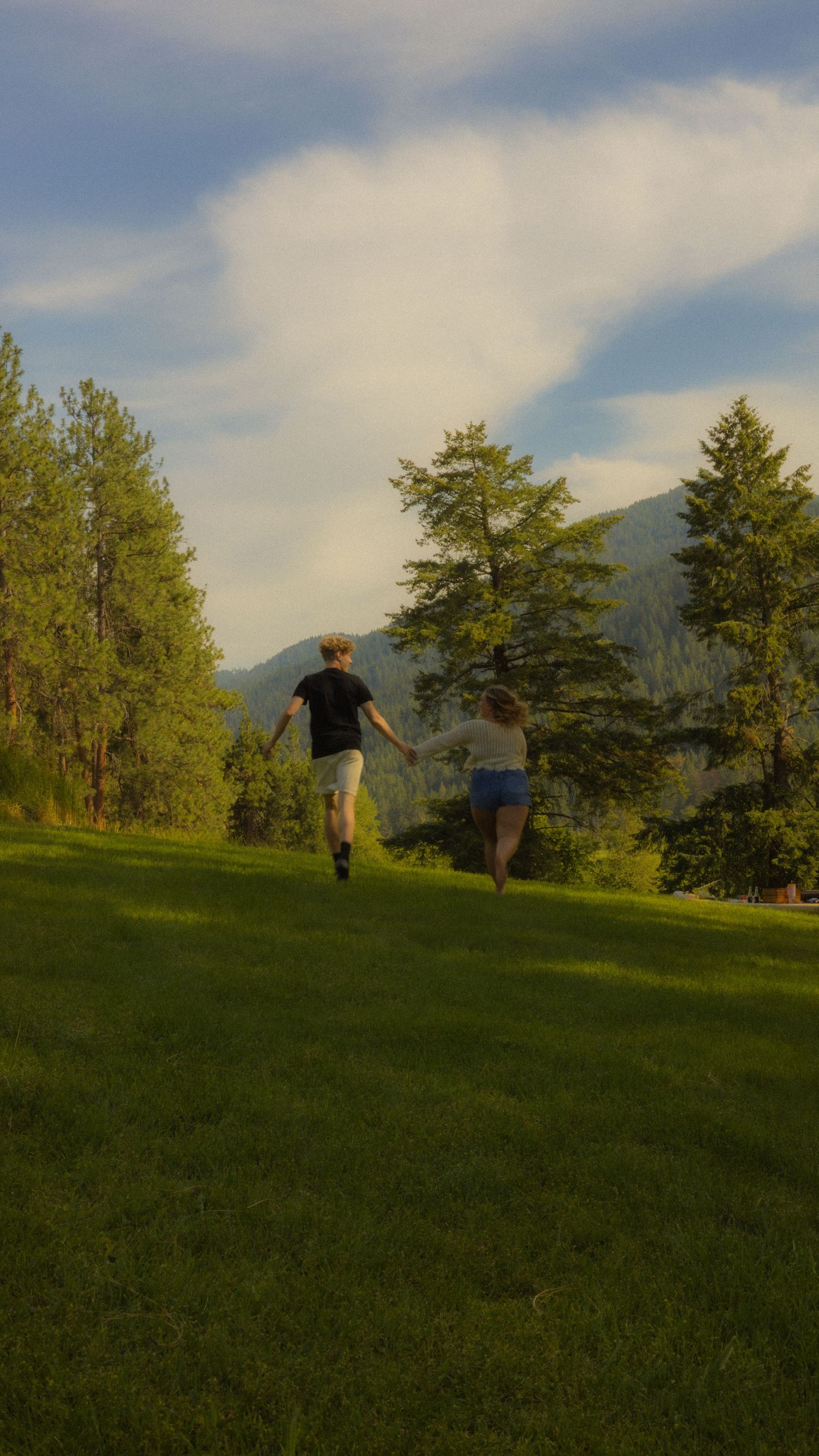 A couple holds hands while running across a lush green hillside with trees and mountains under a partly cloudy sky.