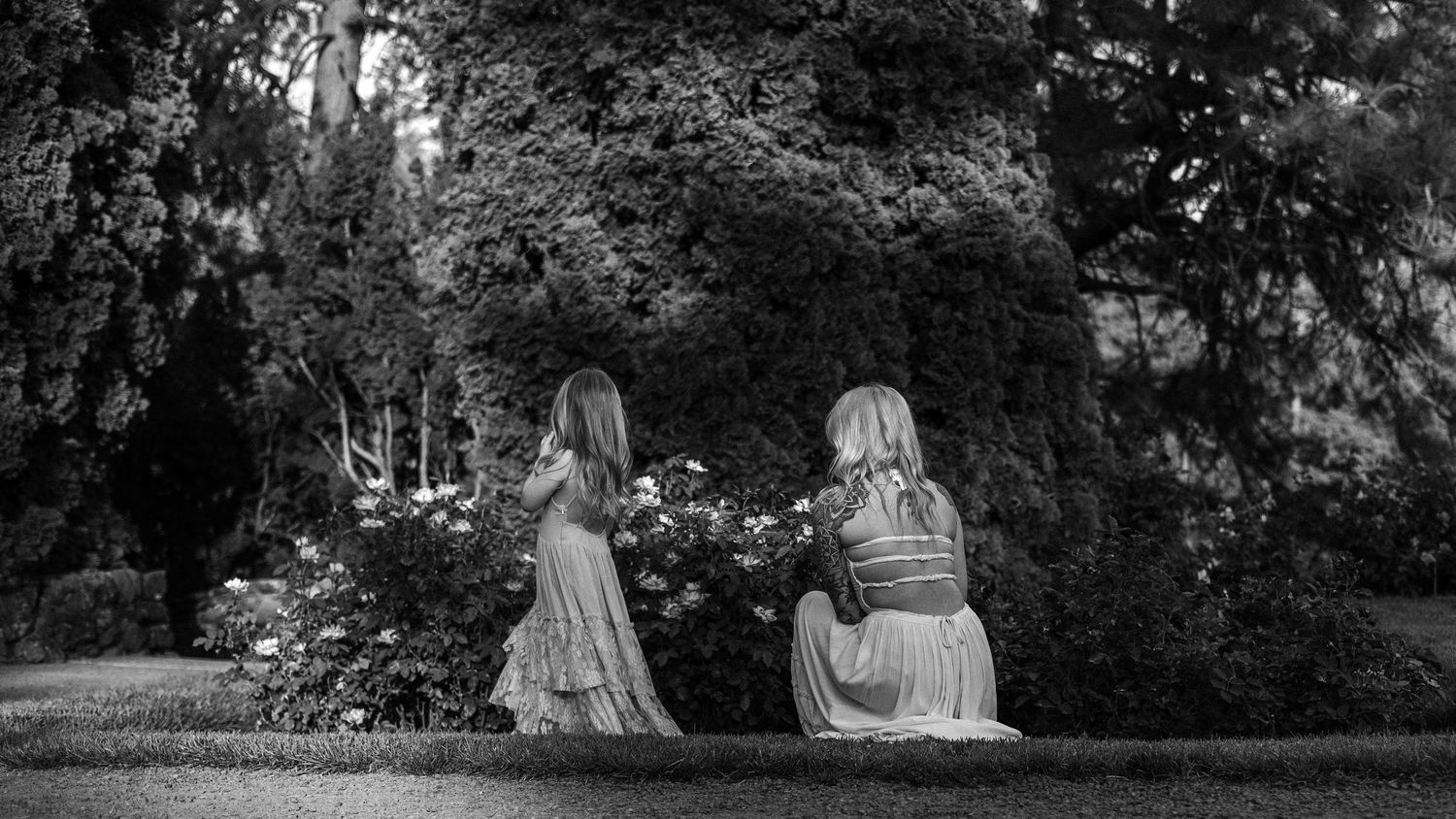 Two girls in dresses explore a lush garden among blooming flowers and tall trees in a serene black and white photo.