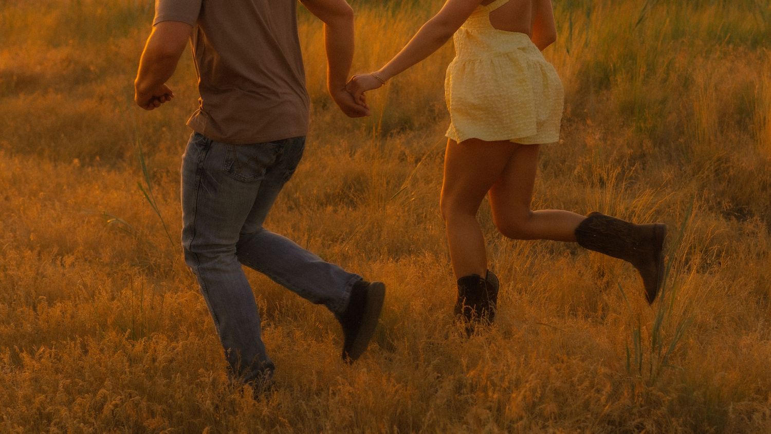 A couple holding hands running through a golden sunlit field at sunset wearing jeans and a white dress.