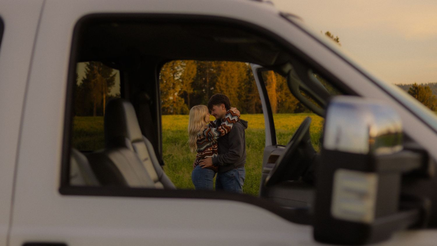 A loving couple embraces in a green field, reflected in the side window of a white truck at sunset.