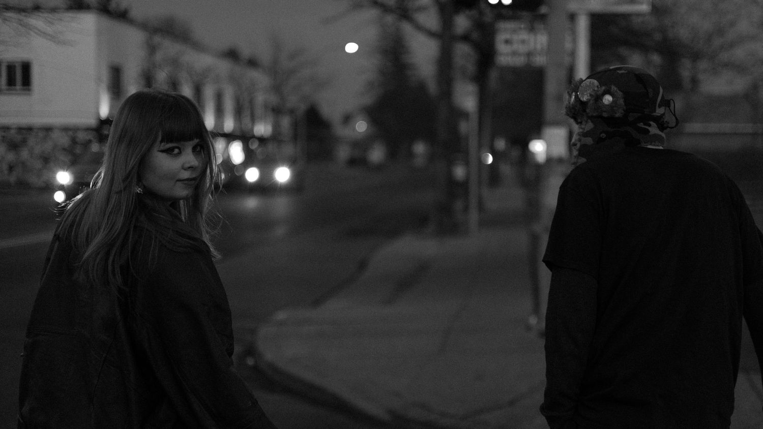 A black and white photo of a woman with long hair and a person wearing a helmet on a quiet street at night.