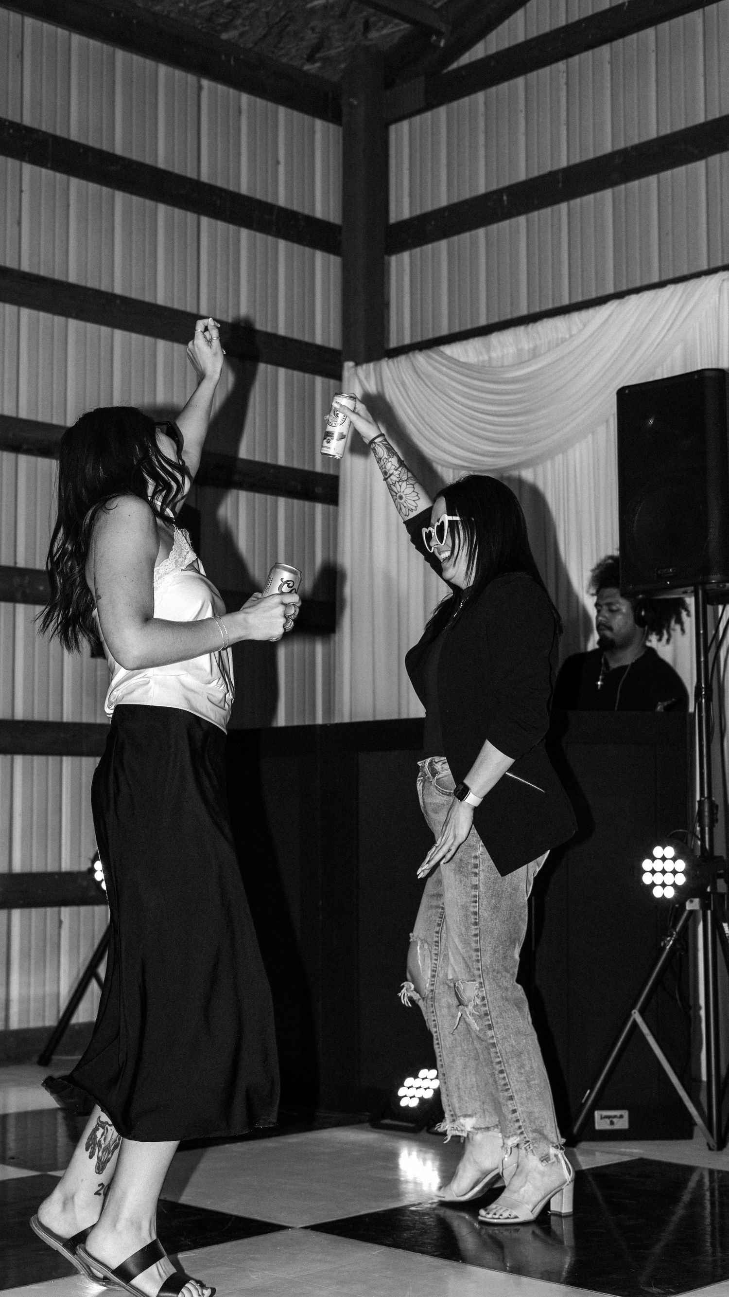 Two women dancing together at a party venue in black and white photo with DJ equipment visible in background.