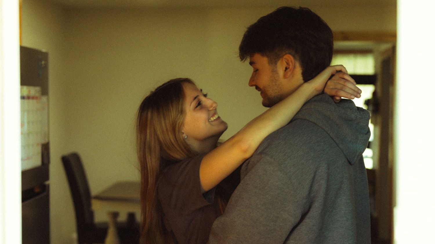 A young couple shares a tender embrace and smile at each other indoors in a warmly lit room.