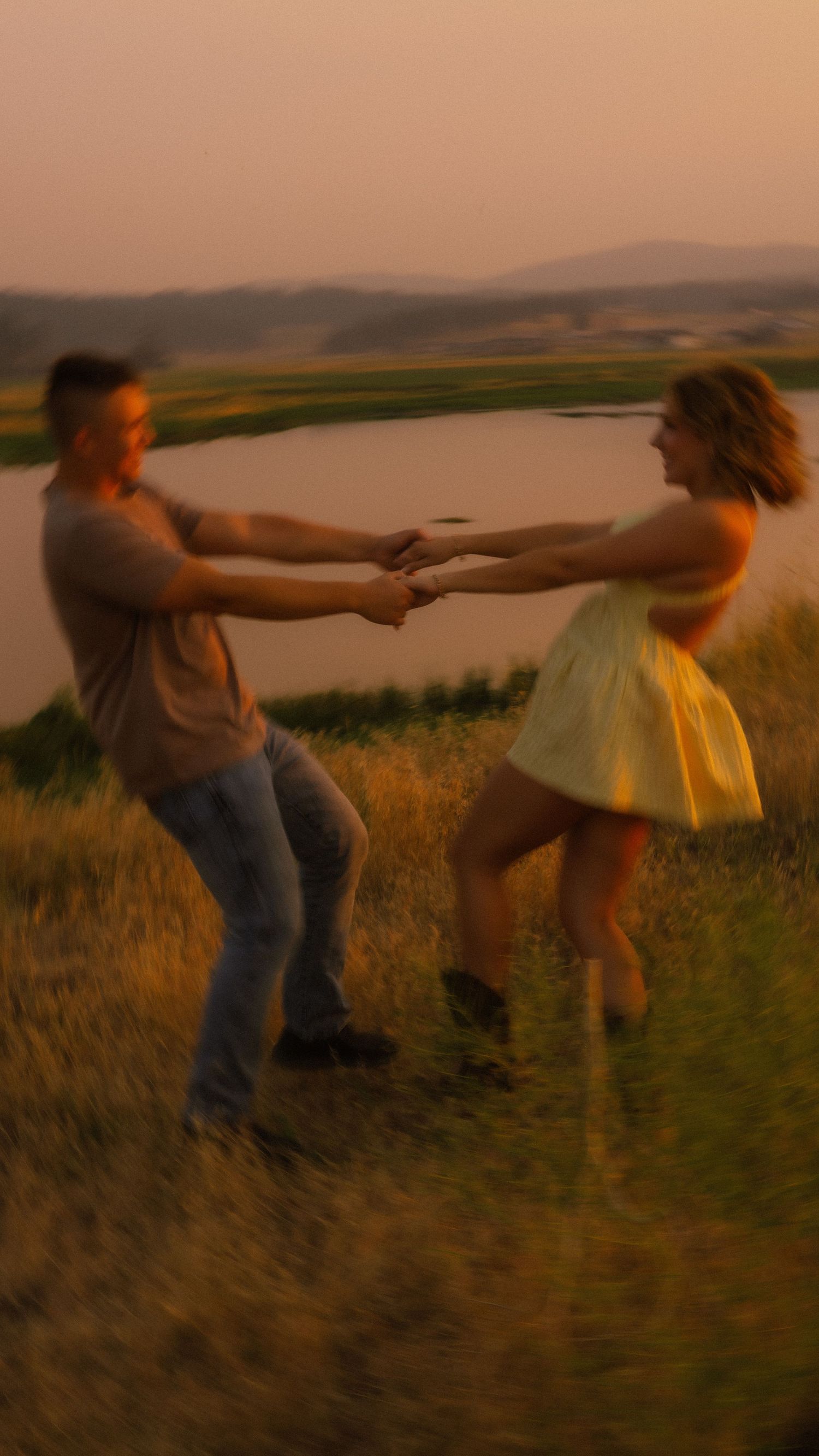Couple dancing joyfully at sunset in a golden meadow near a lake, holding hands and spinning together.