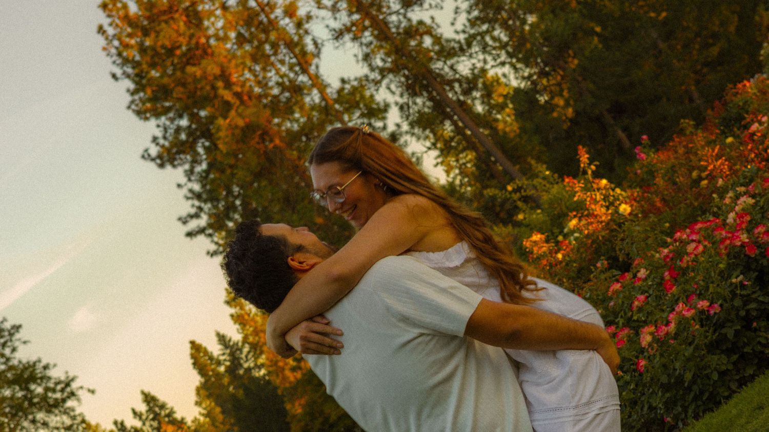 A couple shares a romantic embrace outdoors, with the man lifting the woman among colorful flowers and lush green trees at sunset.
