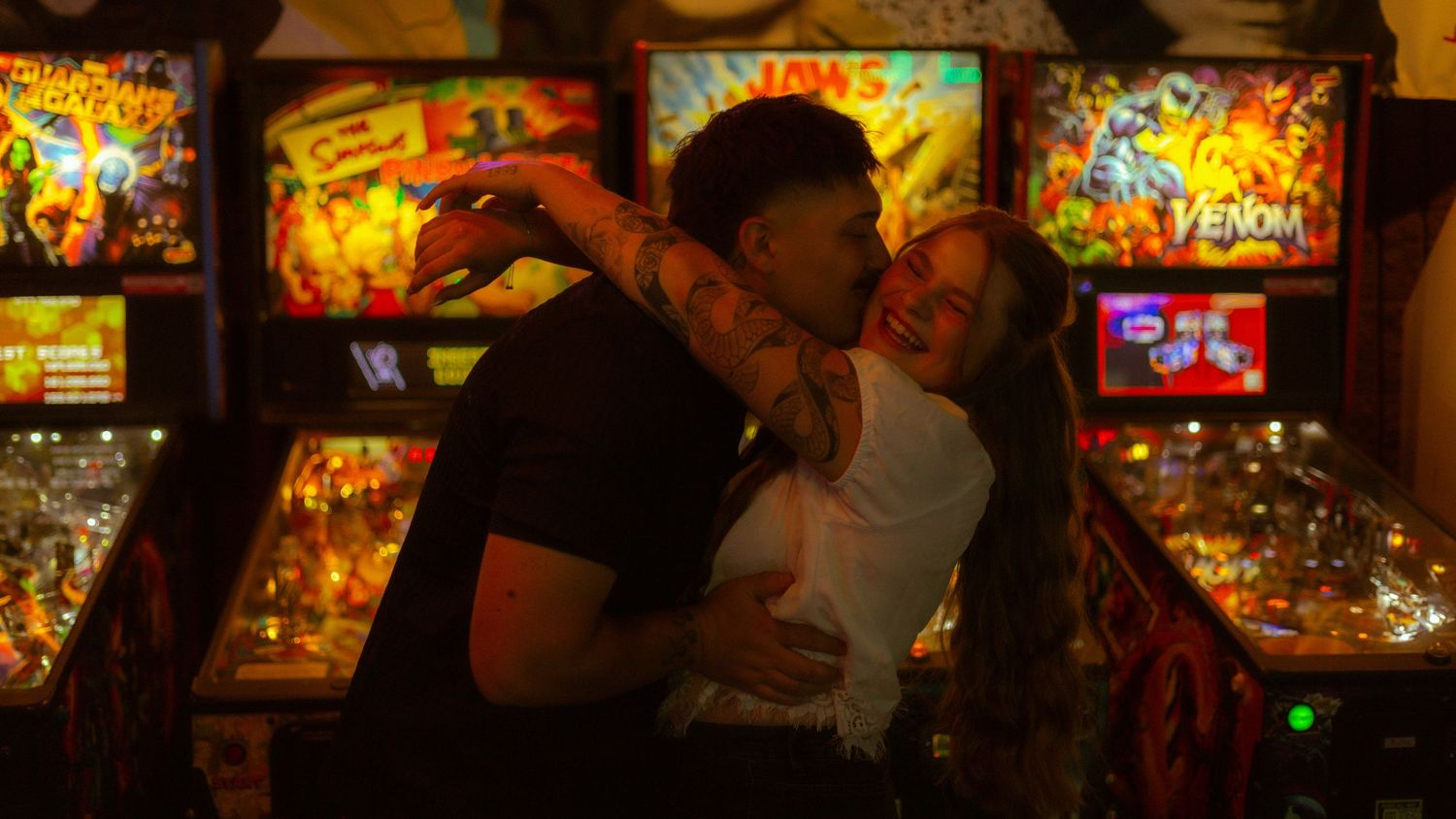 Couple embracing and hugging in front of colorful vintage pinball machines including Venom arcade game.