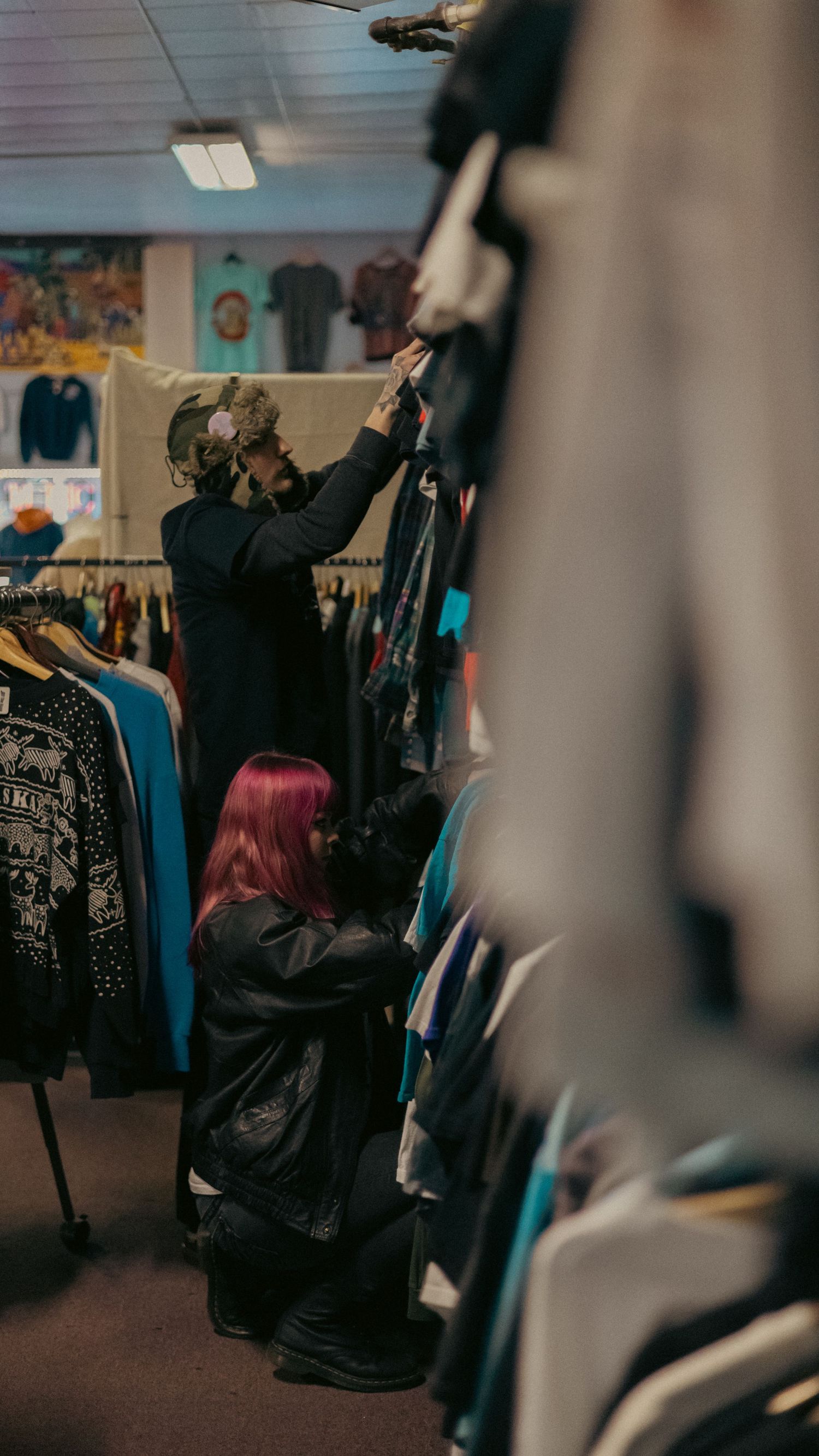 Two people browse colorful clothing racks in a vintage thrift store, one with pink hair crouching below.