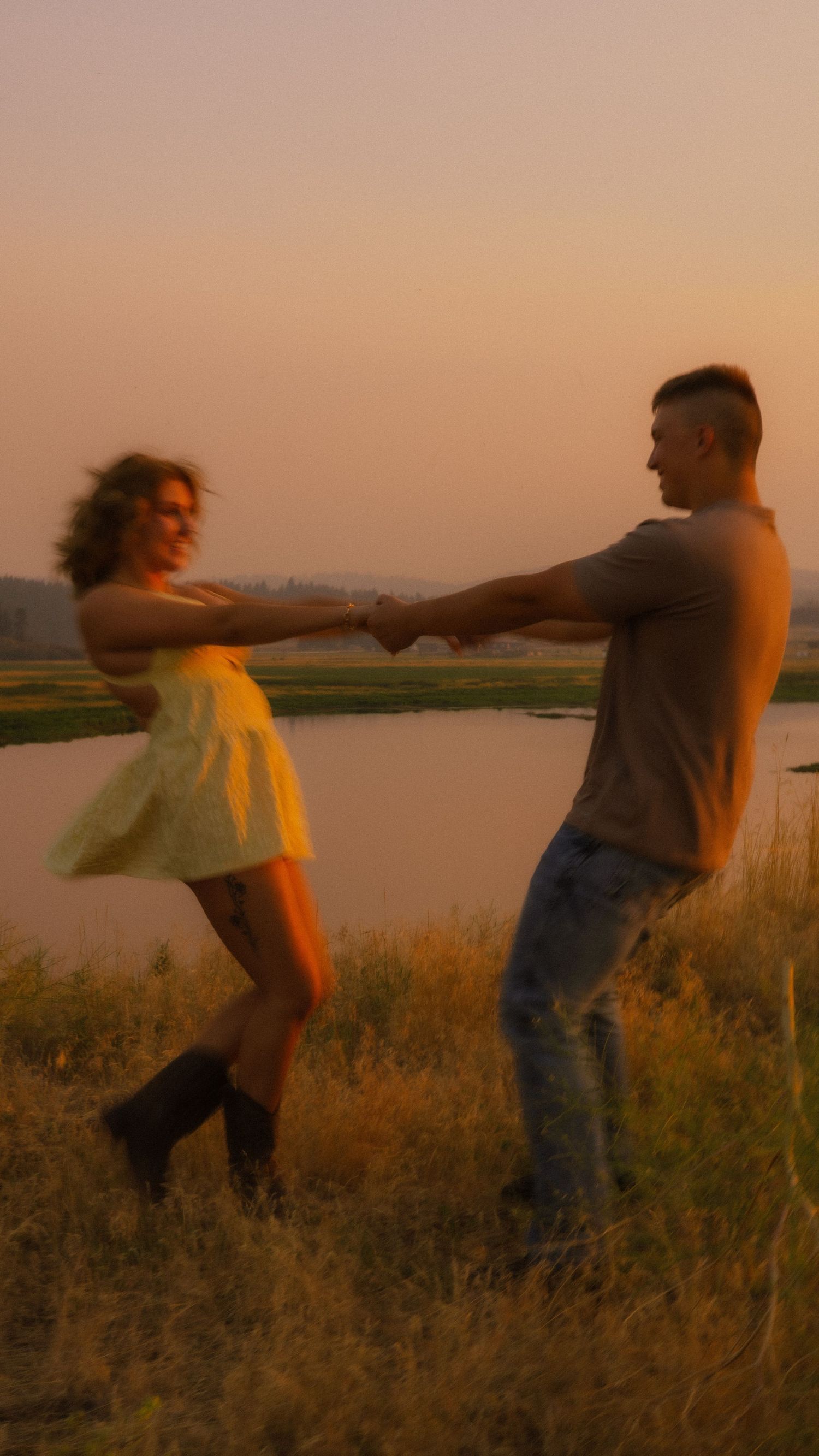 Couple dancing joyfully at sunset by a lake, woman in yellow dress and boots, man in jeans, golden hour glow.