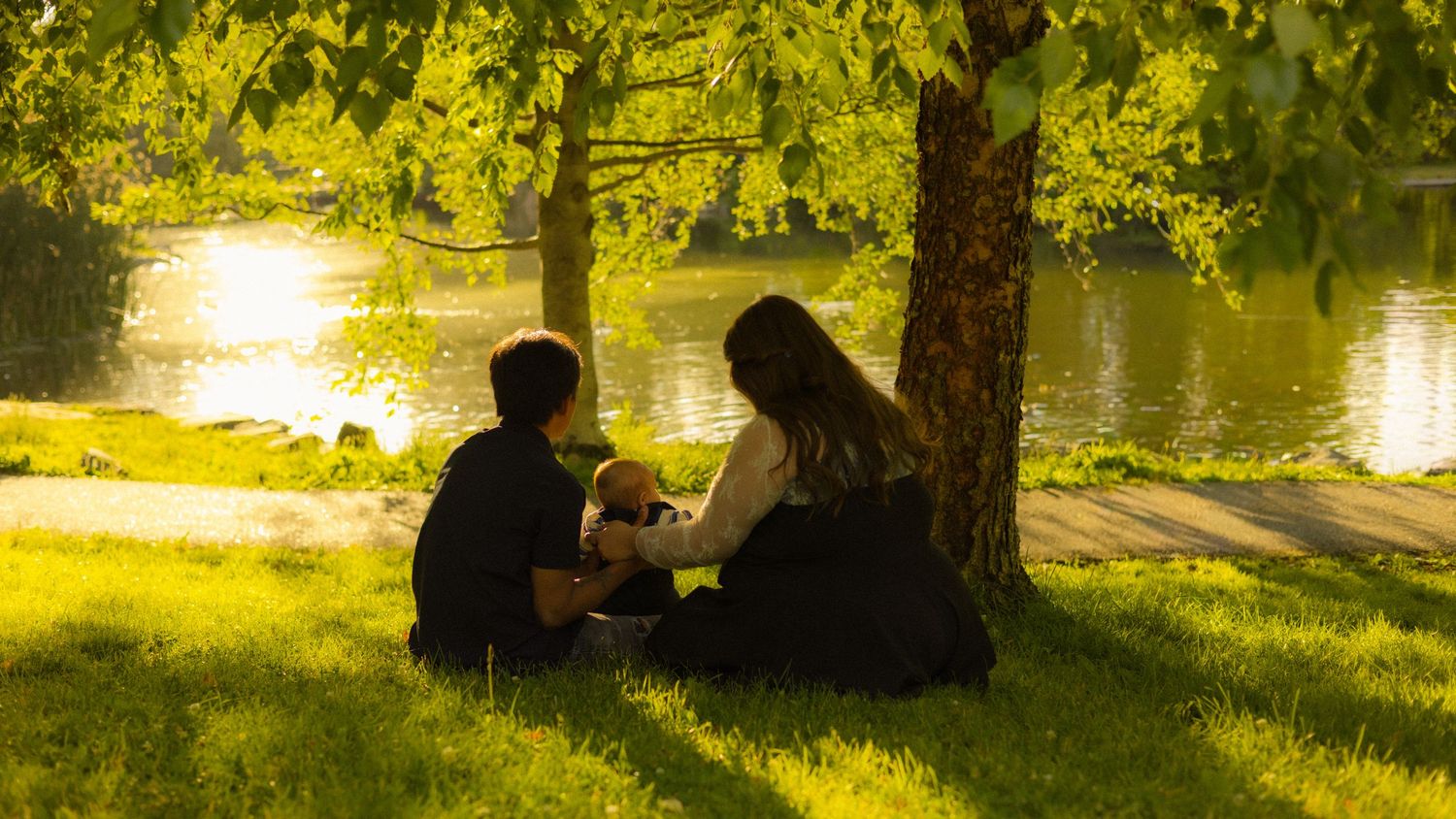 Couple sitting on green grass under a tree by a sunlit river in a peaceful park setting.