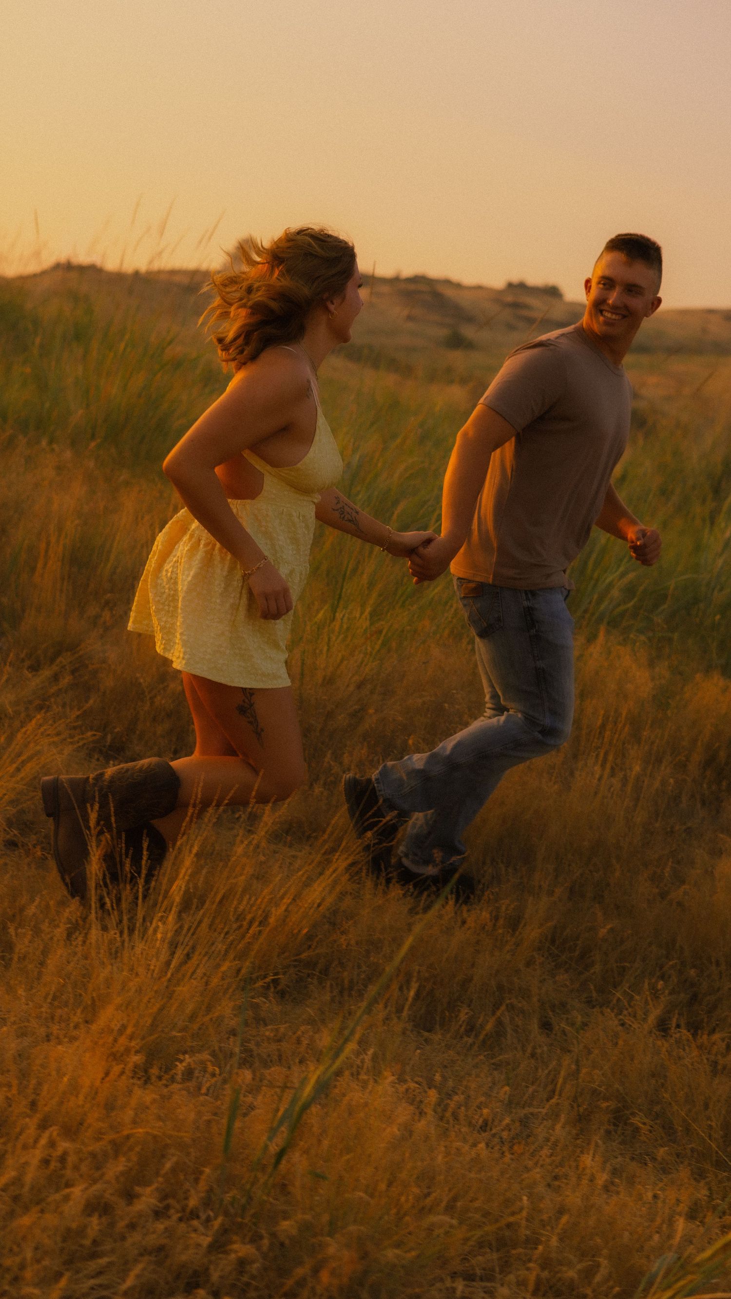 A joyful couple holds hands running through golden grass fields at sunset, woman in yellow dress, man in jeans.