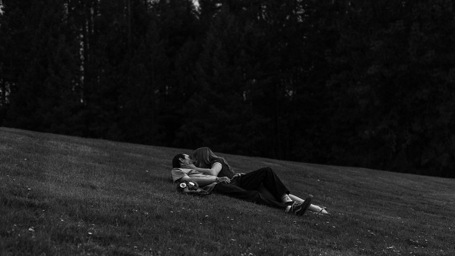Black and white image of a person reclining on a grassy slope with dark trees behind.