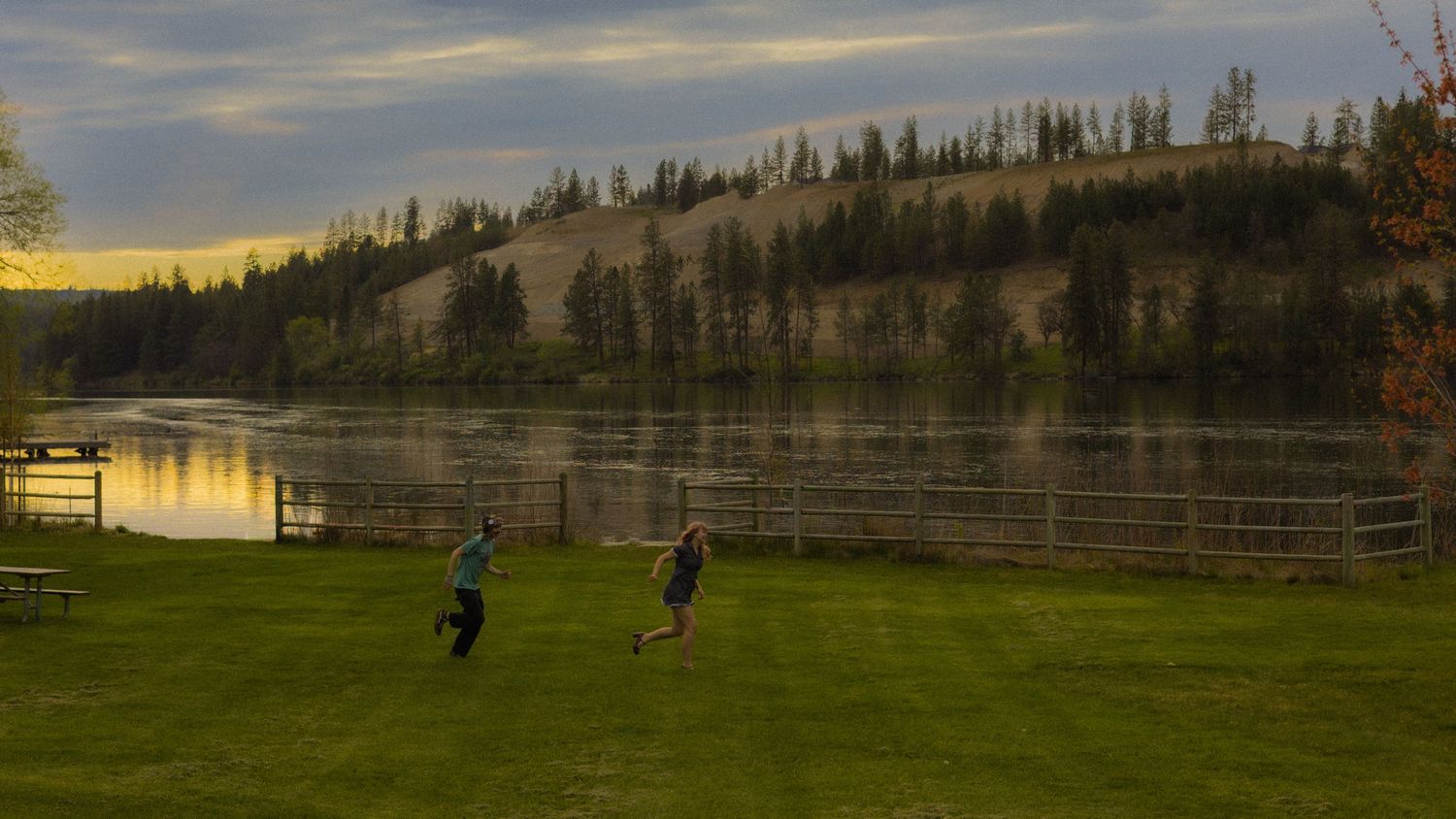 Two people playing frisbee on a green lawn beside a river at sunset in autumn.