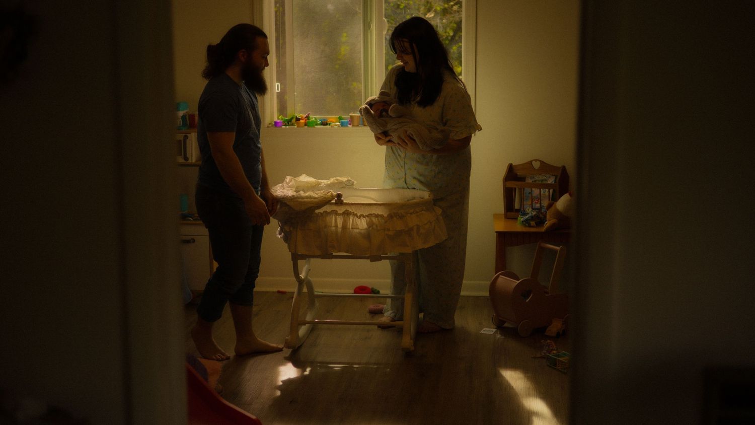 Parents stand together beside a bassinet in a softly lit nursery room holding their newborn baby.