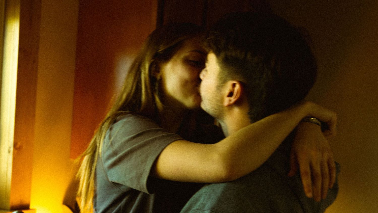 A young couple sharing a romantic kiss in a warmly lit room with amber tones.